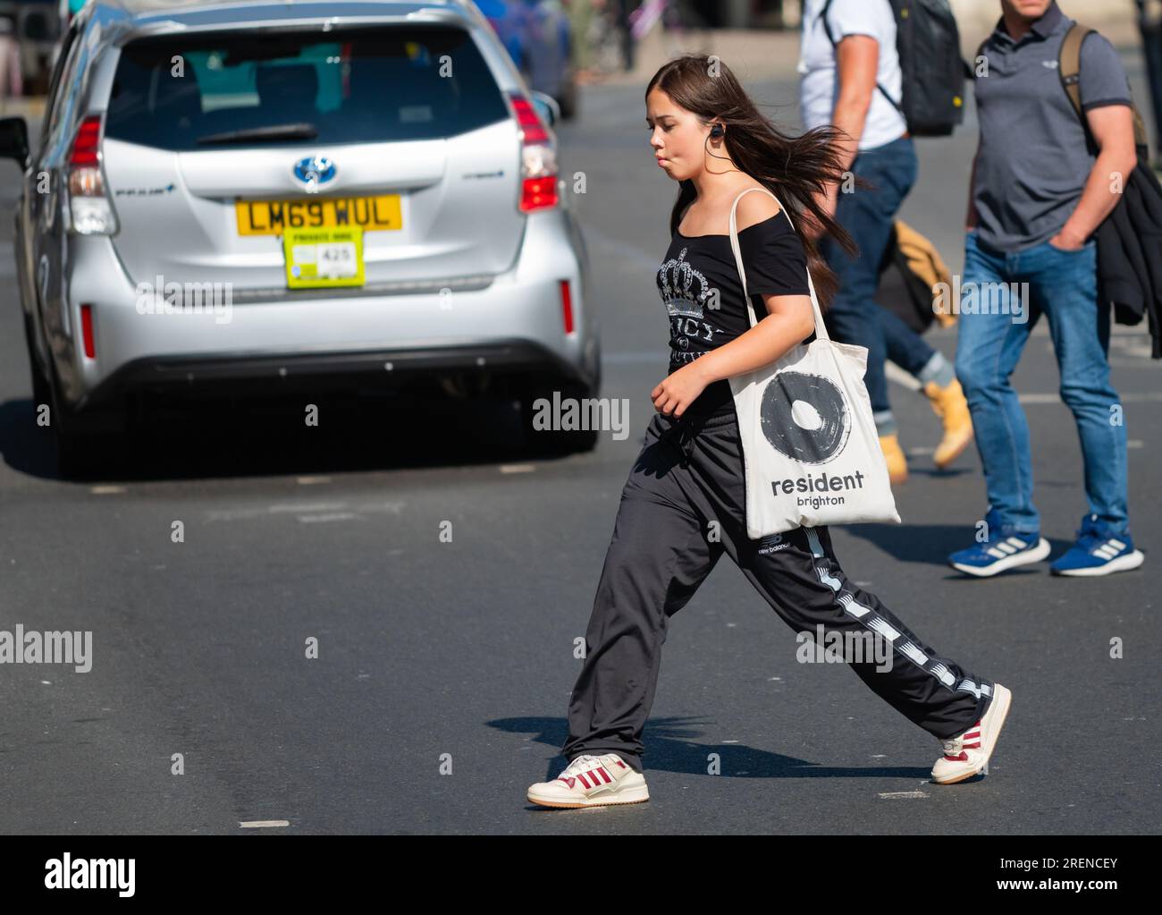 Hübsche junge weiße Frau mit Kopfhörern, modisch gekleidet mit langen Haaren und Schultertasche, überquert die geschäftige Straße im Sommer, Großbritannien. Stockfoto