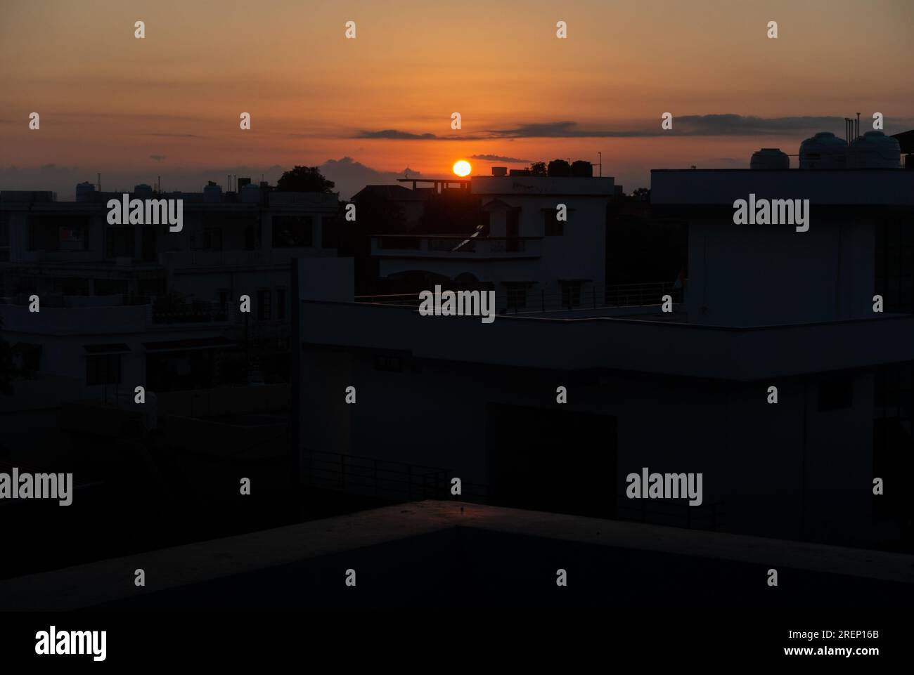 Die Stadtlandschaft in der Dämmerung mit einem orangefarbenen Abendhimmel und einer runden Sonne am Horizont, die von einem Dach in Uttarakhand, Indien, eingefangen wurde. Stockfoto