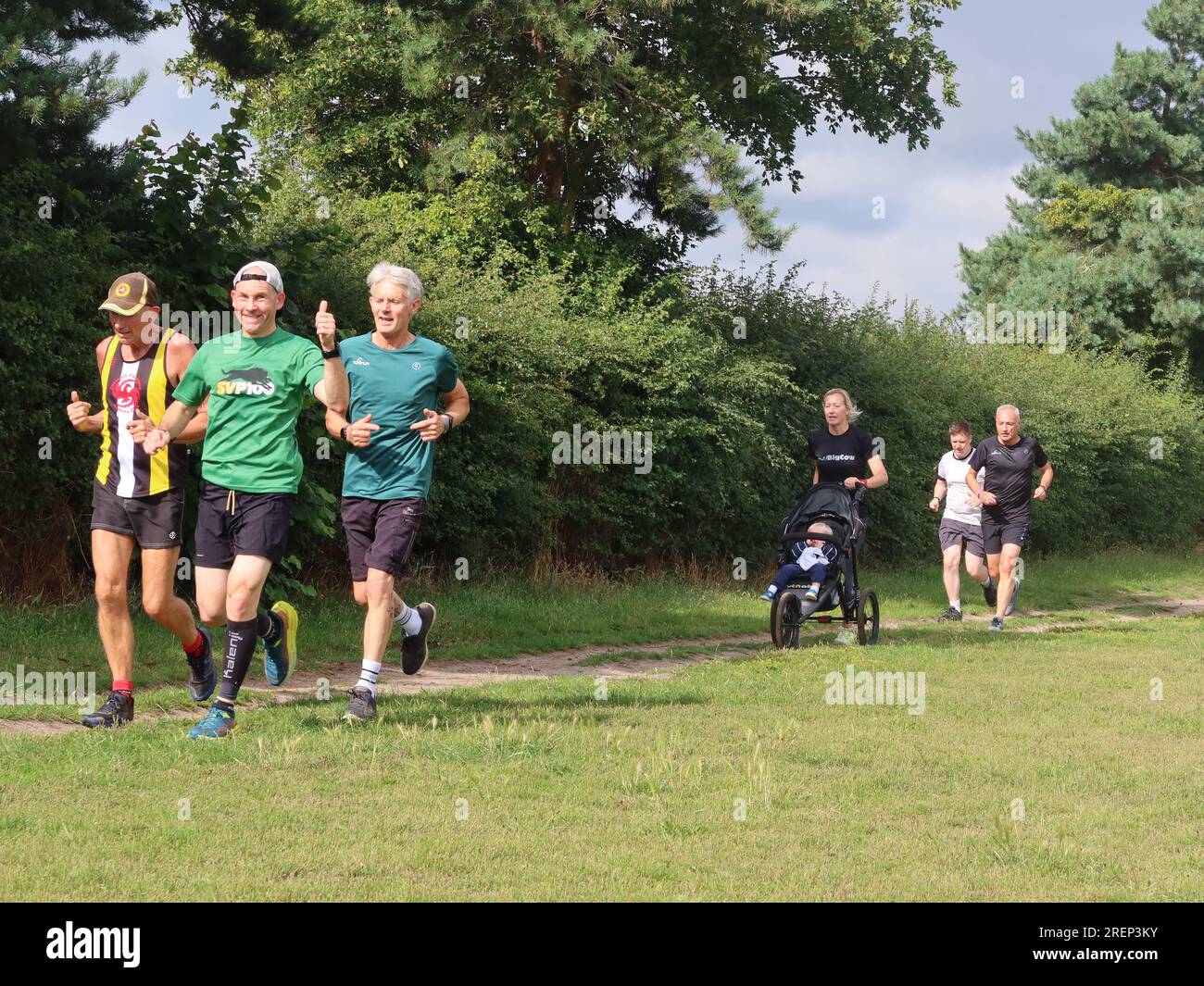 Kesgrave, Suffolk - 29. Juli 2023 : warmer Sommer am Samstagmorgen Parkrun im Millennium Field. Daumen hoch von einem lächelnden Läufer. Der Buggy kommt von hinten. Stockfoto