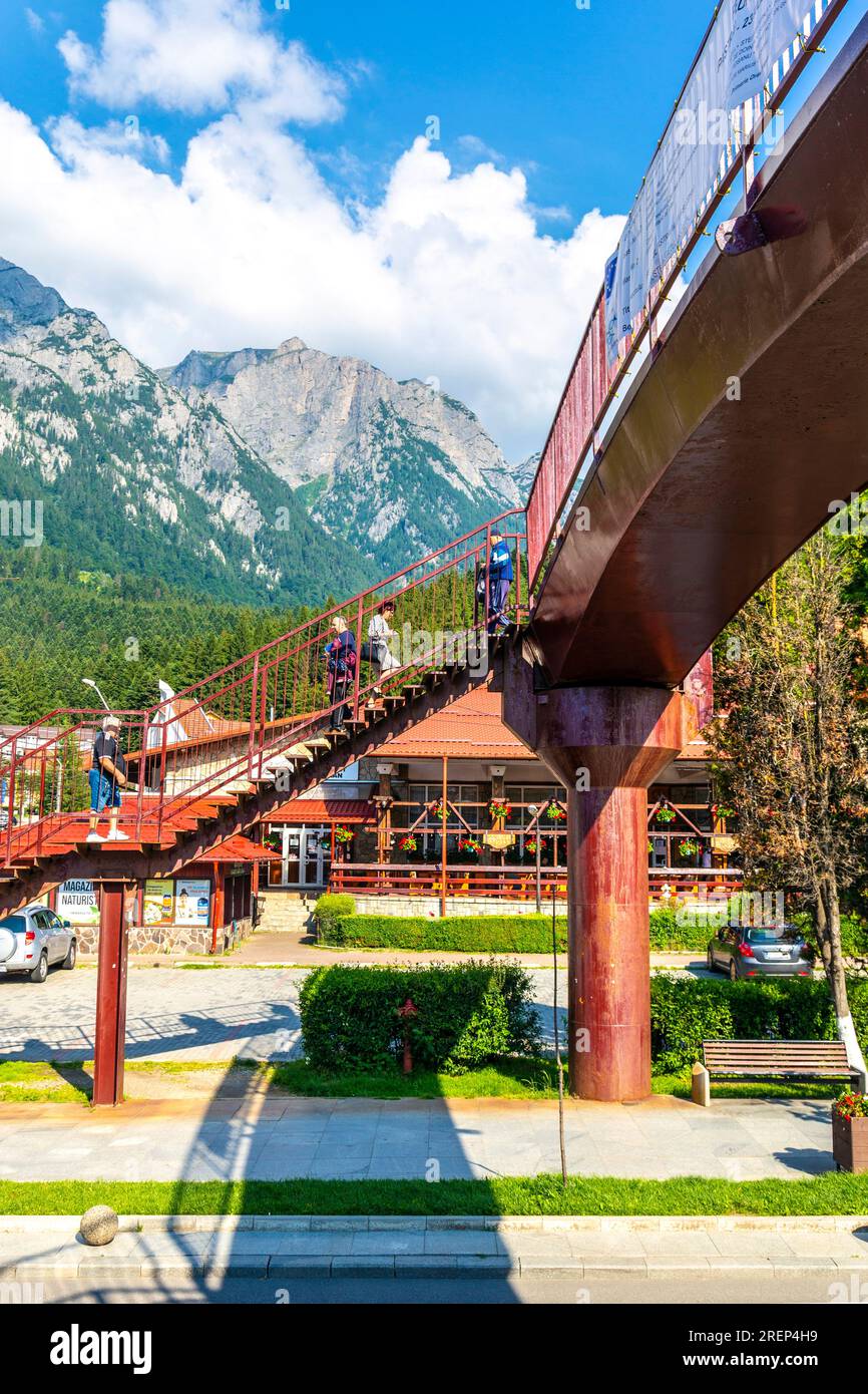 Menschen überqueren die rote Fußgängerbrücke über die Hauptstraße Bulevardul Libertății und das Bucegi-Gebirge, Busteni, Rumänien Stockfoto