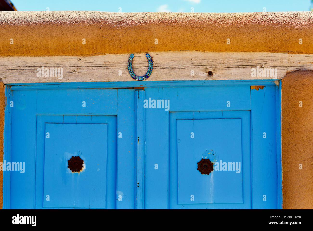 Ranchos de Taos, New Mexico, USA - 23. Juli 2023:Ein dekoriertes Hufeisen hängt über den blau bemalten Holzdoppeltüren eines traditionellen Hauses aus lehmziegeln. Stockfoto