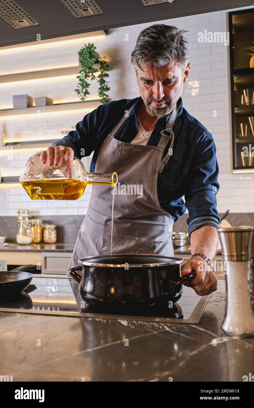 Fokussierter, bärtiger Koch in einer Schürze, der Öl aus der Flasche in den Topf gießt, während er in einer hellen, modernen Küche kocht Stockfoto