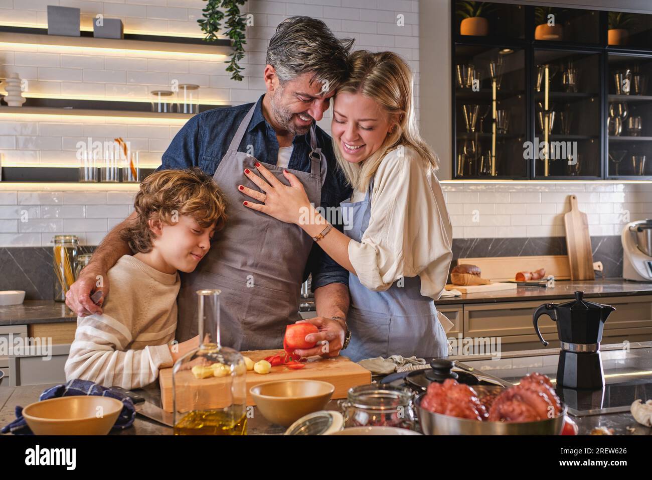 Fröhlicher Mann mit Tomate in der Hand, umarmte Frau und Sohn, während er zusammen in der Küche Krake kochte Stockfoto
