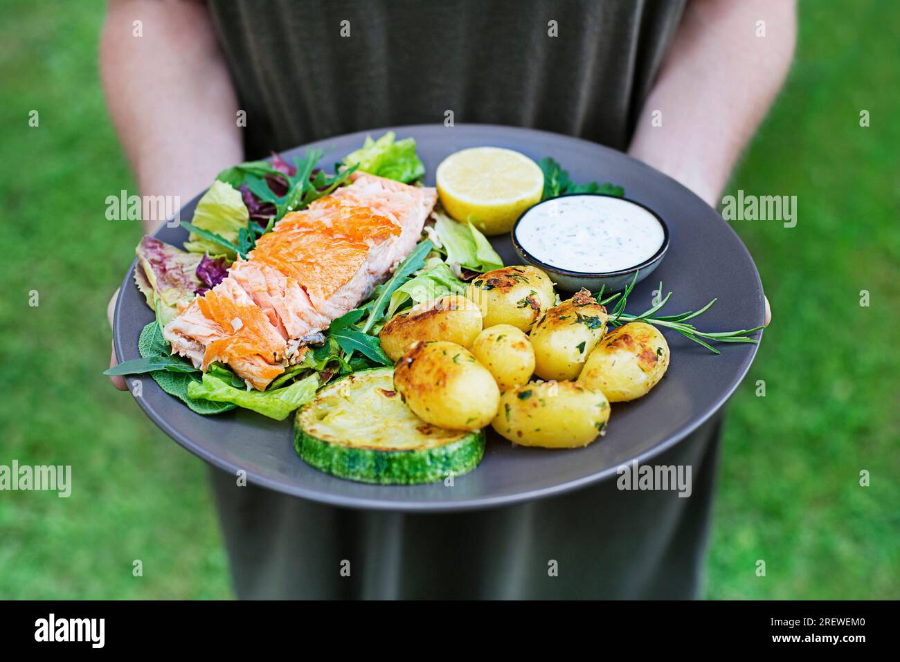 Frauenhände halten gebackenes Lachsfilet mit Kartoffeln und Gemüse in der Gartensaison. Stockfoto