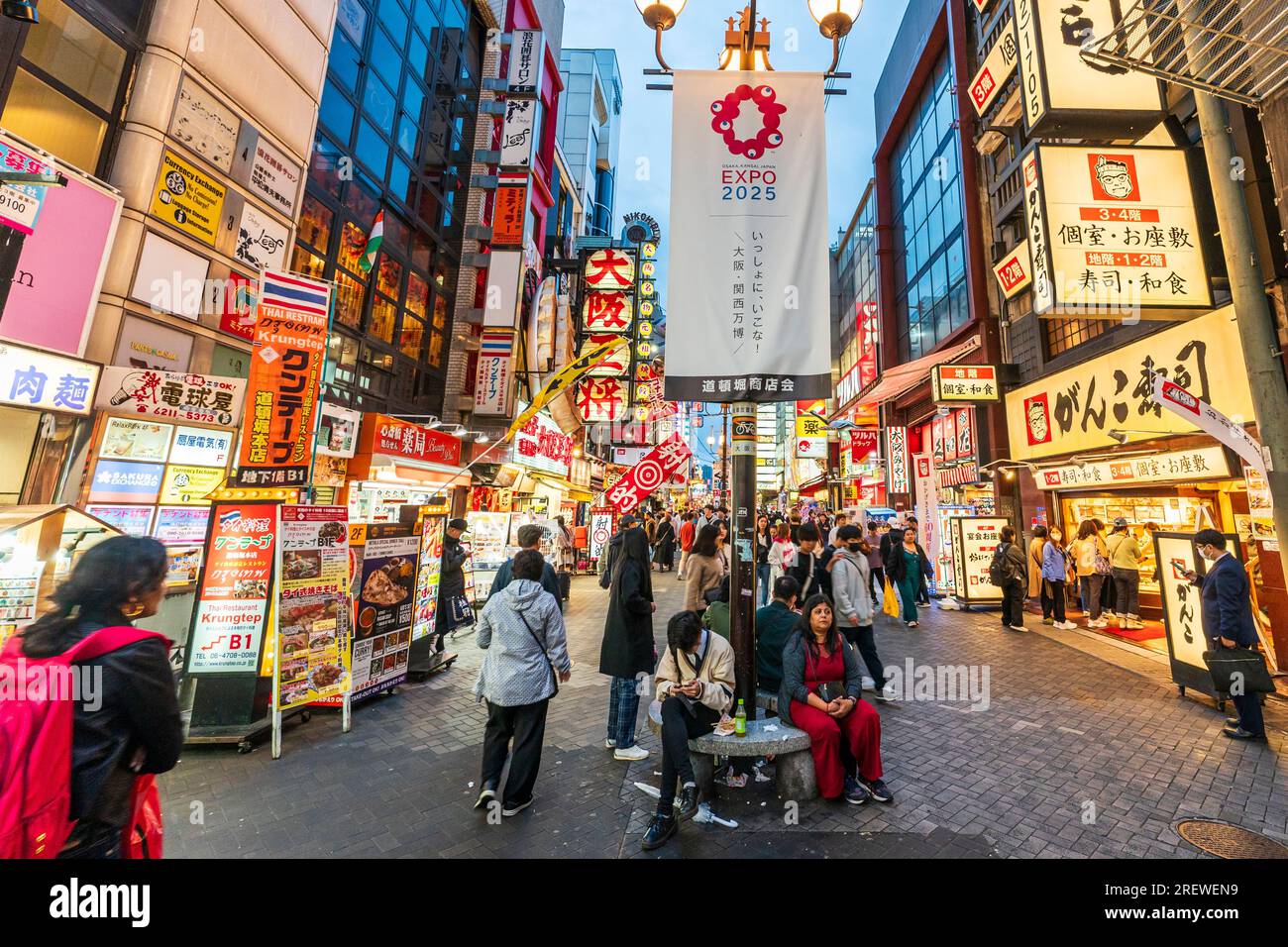 Blick auf Dotonbori, am frühen Abend, voller Menschen. Straßenbeleuchtungsstand im Vordergrund mit expo 2025-Banner, hinter den vielen Neonschildern. Stockfoto