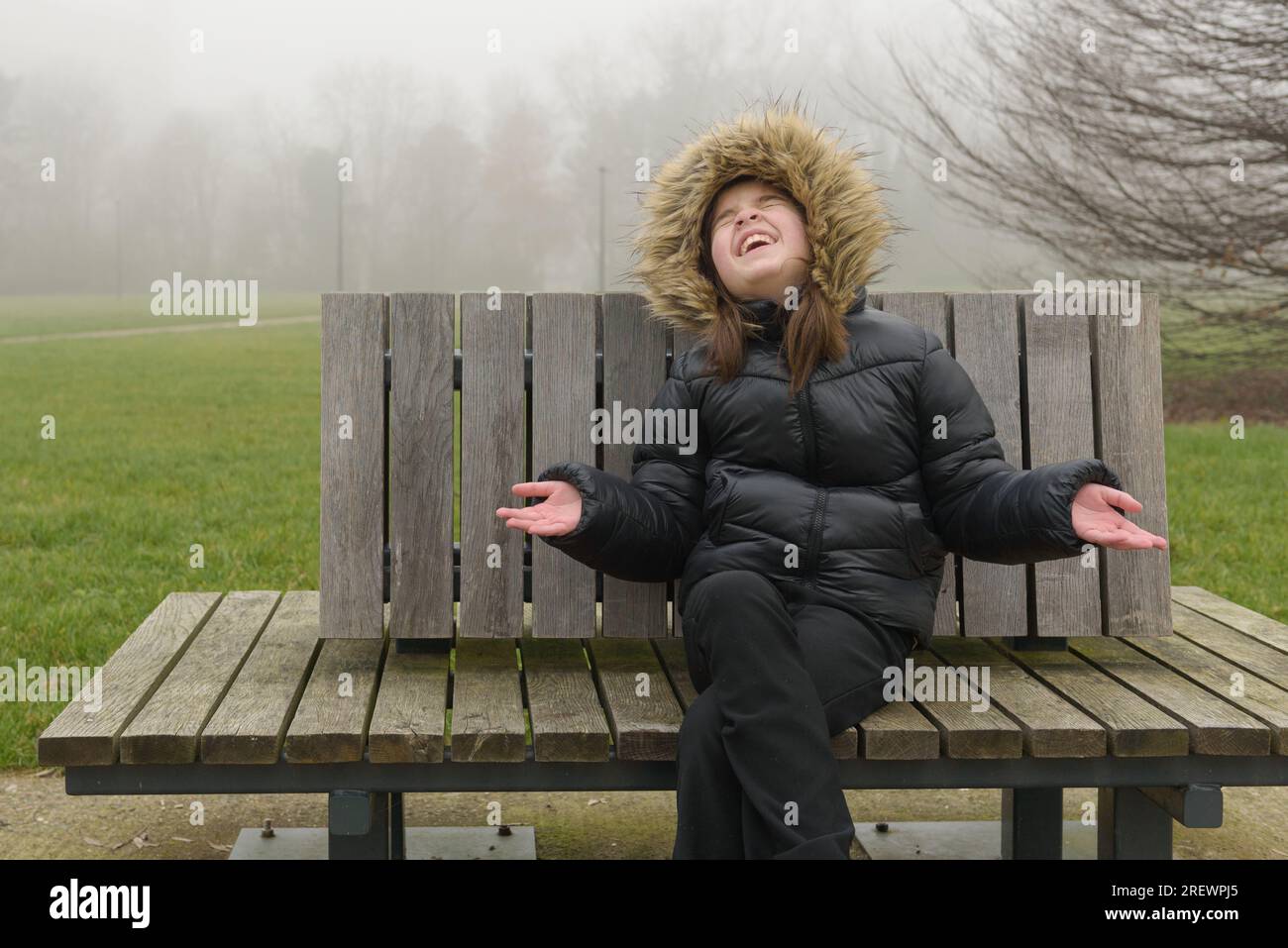 Ein Mädchen mit guter Laune auf einer Parkbank im Winter. Warme Jacke, Nebel Stockfoto