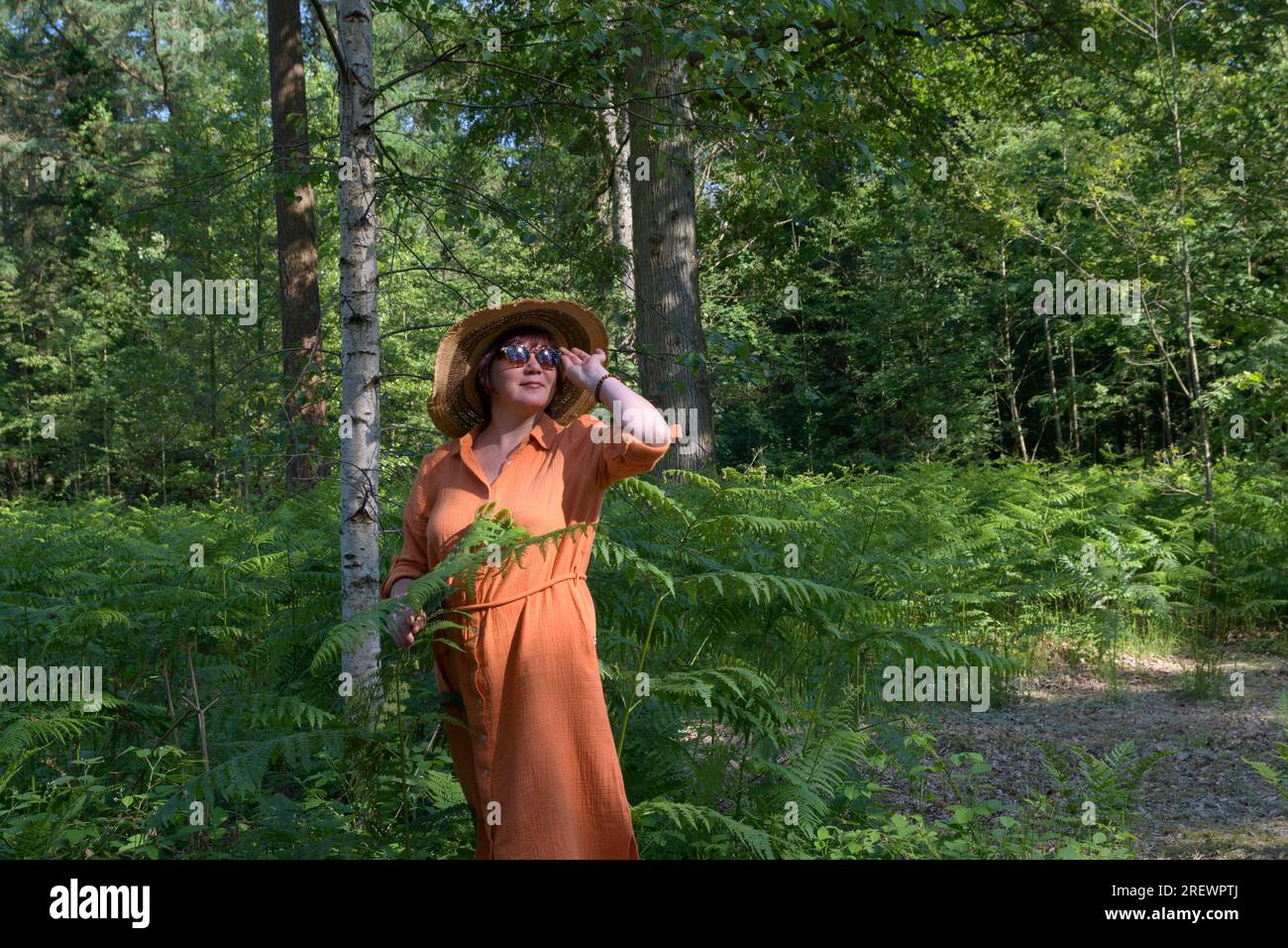 Eine Frau mittleren Alters in einem schattigen grünen Park. Orangefarbenes Kleid, Strohhut, Sonnenbrille. Platz für Text Stockfoto