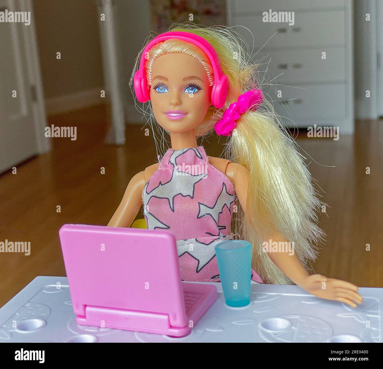 Die Plastik-Barbie-Puppe sitzt mit einem Laptop und einer Tasse an einem Tisch und hört Musik über ihre Kopfhörer. Stockfoto