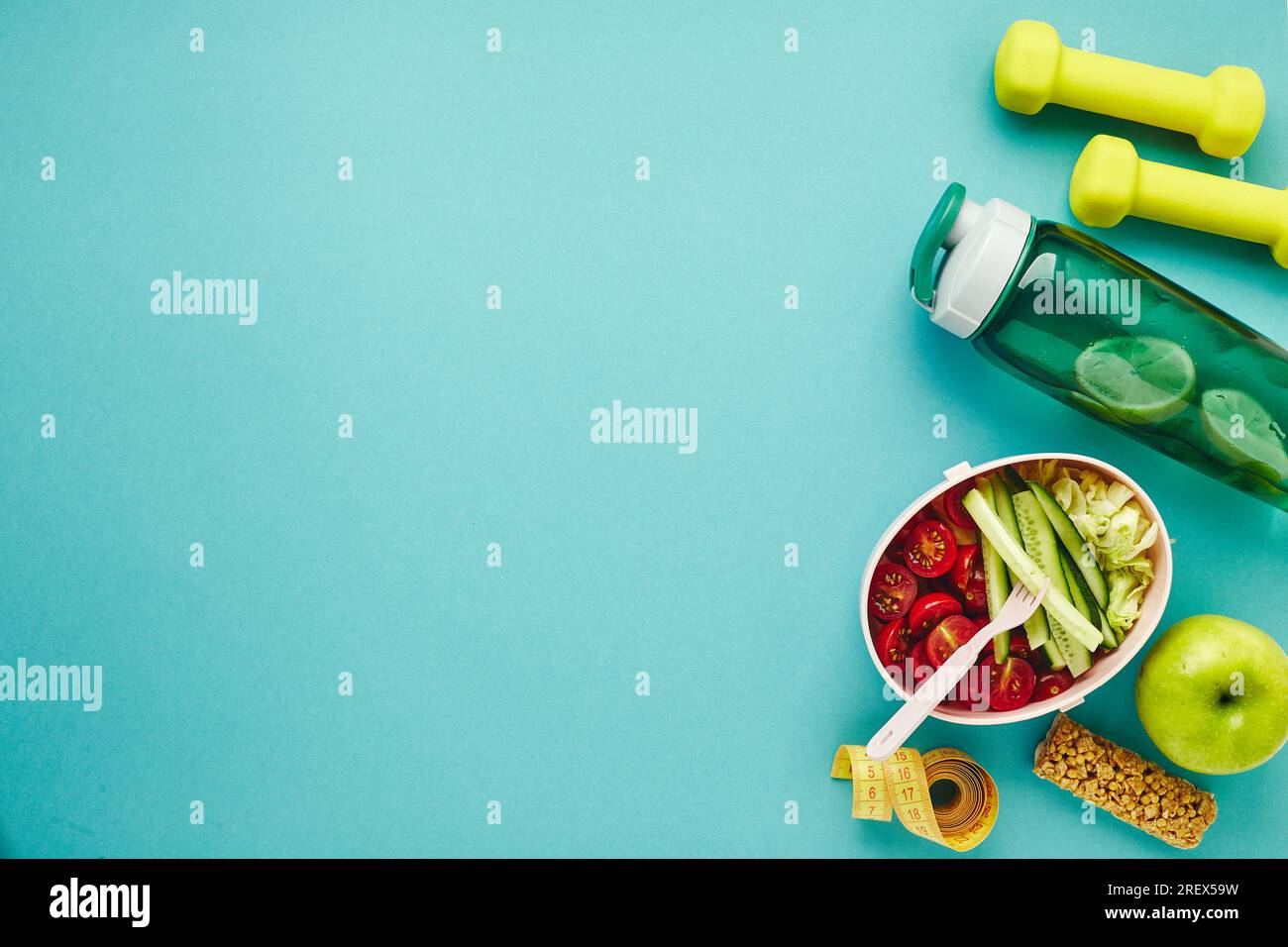 Kreative Planlage von Sport- und Fitnessgeräten. Kurzhanteln, Maßband, Flasche Wasser und Lunchbox mit gesundem Gemüsesalat auf hellblau Stockfoto