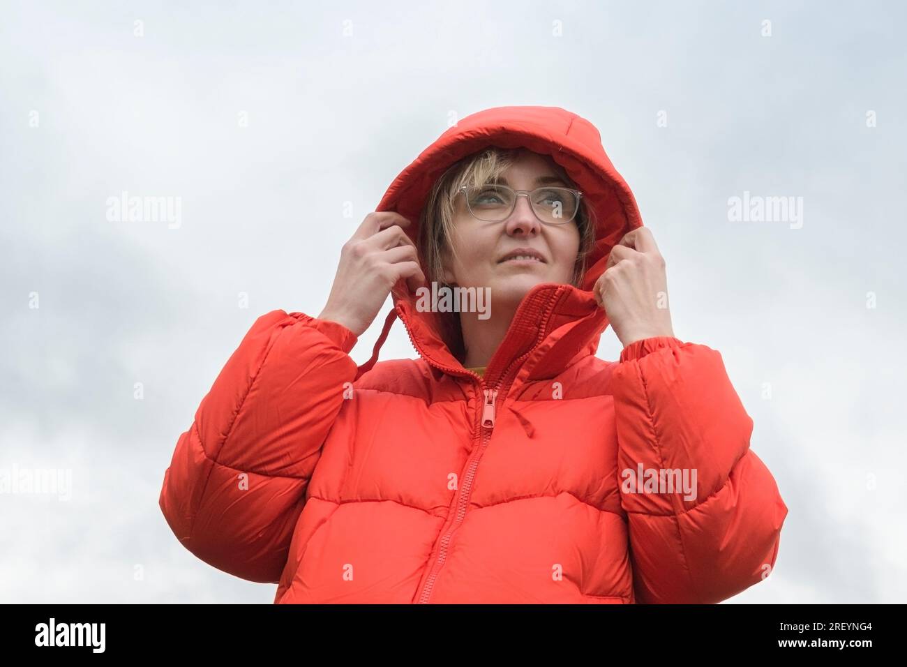 Porträt einer Frau in einer roten Jacke mit Kapuze auf dem Kopf vor einem bewölkten Himmel Stockfoto