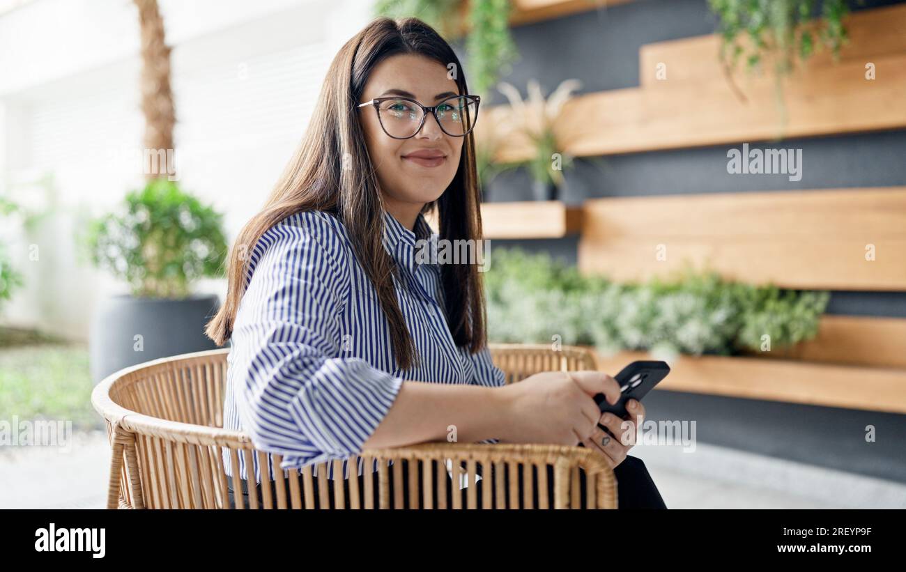 Junge, wunderschöne hispanische Frau, die mit ihrem Smartphone lächelt, sitzt auf einem Stuhl im Bürogarten Stockfoto