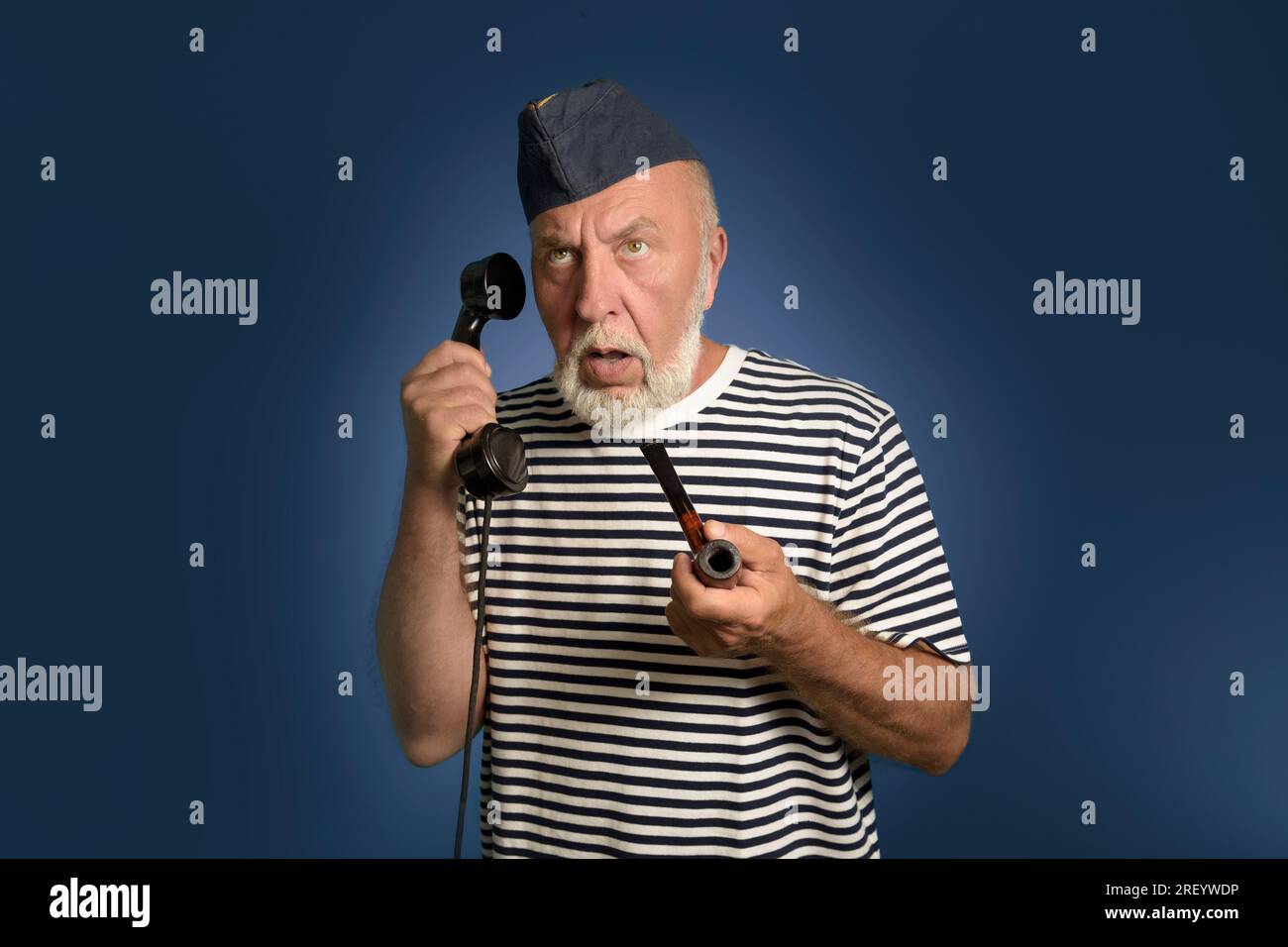 Ein echter Seemann aus der Vergangenheit spricht am Telefon. Hochformat auf blauem Hintergrund. Ein alter Seemann mit einer Rauchpfeife und einem alten Telefon in Hi Stockfoto
