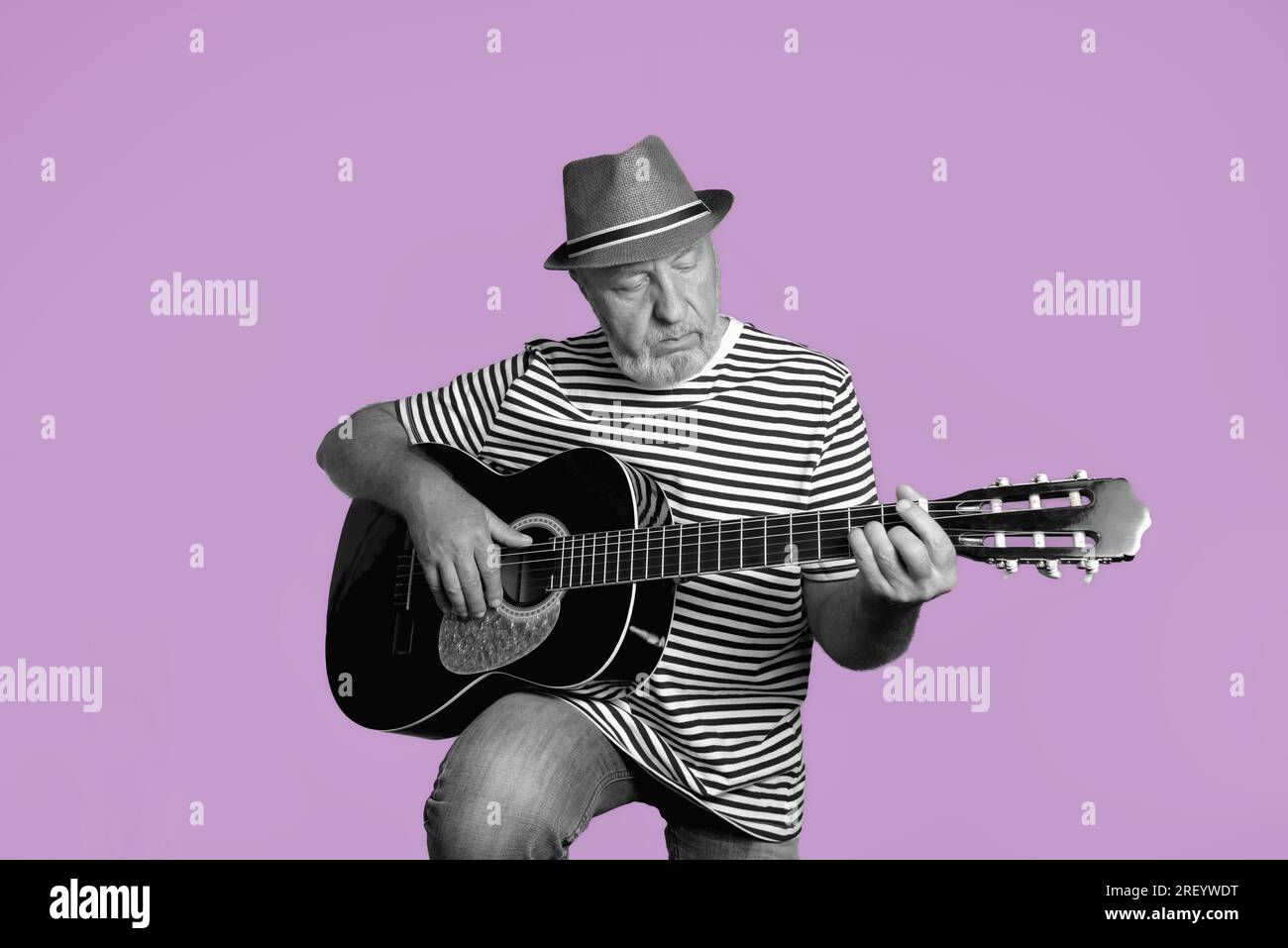 Ein älterer Mann spielt Akustikgitarre. Gestreiftes T-Shirt, Strohhut auf dunkelrosa Hintergrund. Retro-Konzept. Schwarzweiß-Porträt auf dunkelrosa Stockfoto