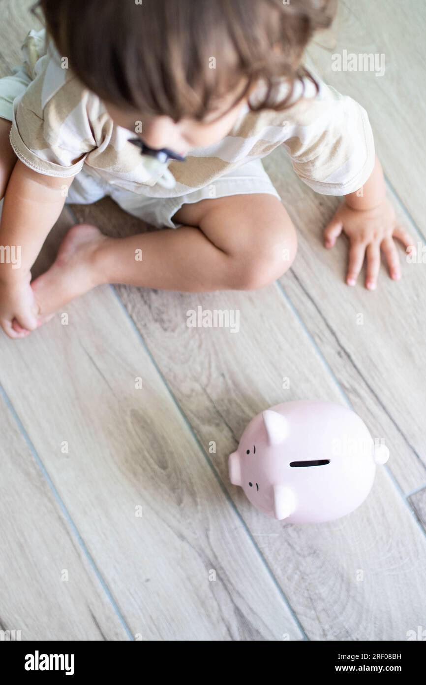 Weißes Kleinkind mit Herzschrittmacher, das auf dem Boden neben einem lächelnden Sparschwein sitzt. Das Sparkonzept für Kinder. Vertikale Aufnahme, Draufsicht. Stockfoto