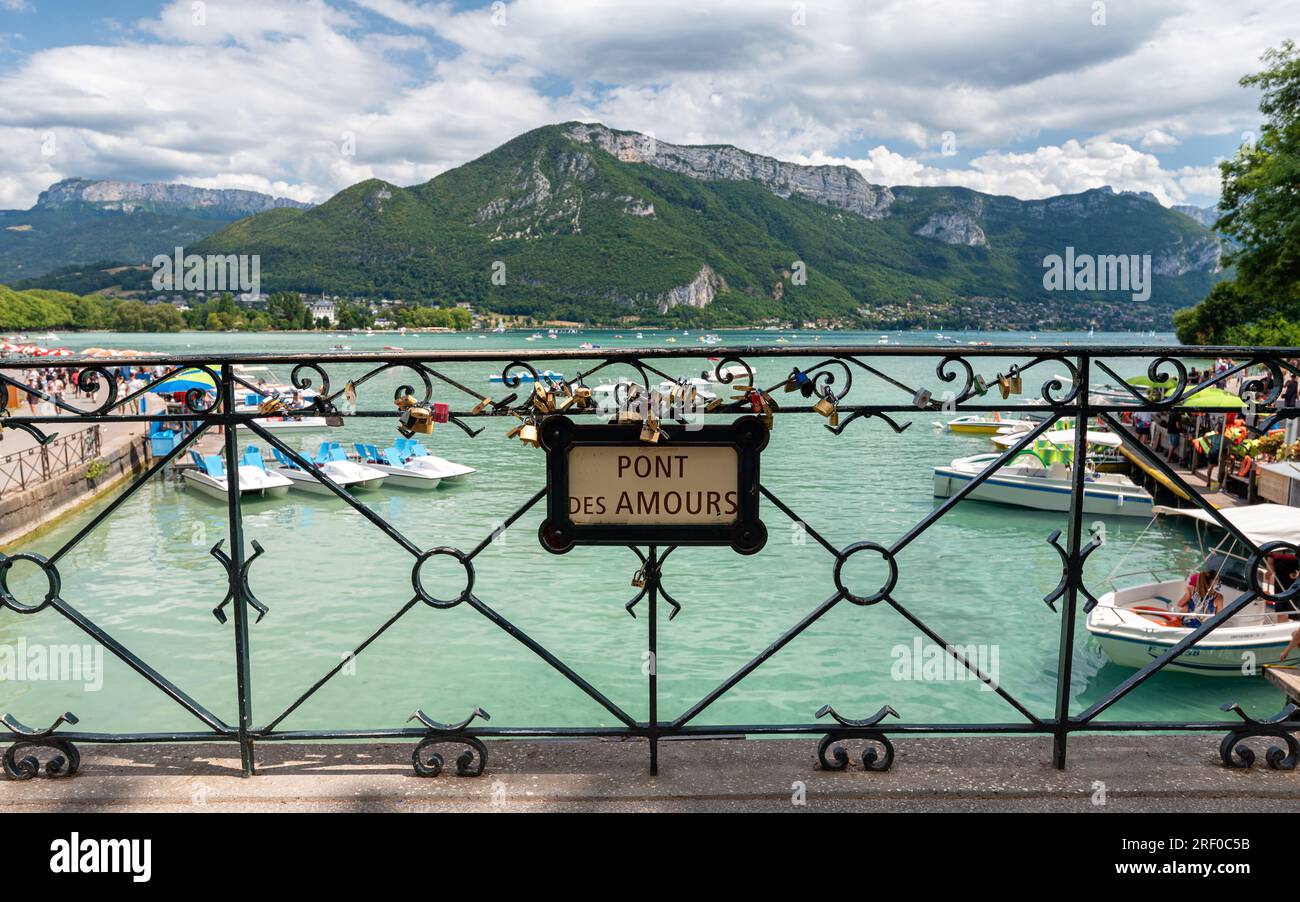 Bridge of Love („Pont des Amours“ auf Französisch auf dem Schild) mit Freizeitaktivitäten und Booten, die auf dem Lake Annecy im Hintergrund segeln. Stockfoto