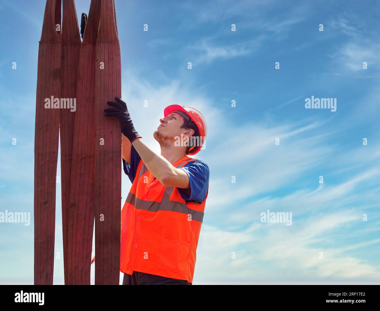 Porträt eines jungen kaukasischen Schleuders bei der Arbeit gegen den Himmel. Der Baumeister oder Handwerker lässt sich an den Hebegurten einhaken und schaut nach oben. Arbeiter mit Helm und Weste. Stockfoto
