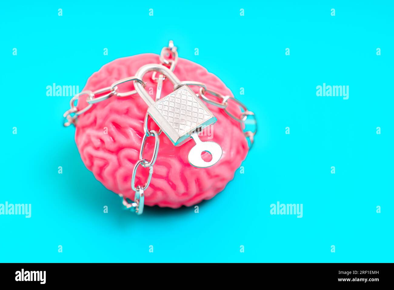 Nahaufnahme eines gelartigen menschlichen Gehirnmodells, das durch Ketten begrenzt und mit einem Vorhängeschloss auf blauem Hintergrund gesichert ist. Der Gedanke, geistig zu sein Stockfoto