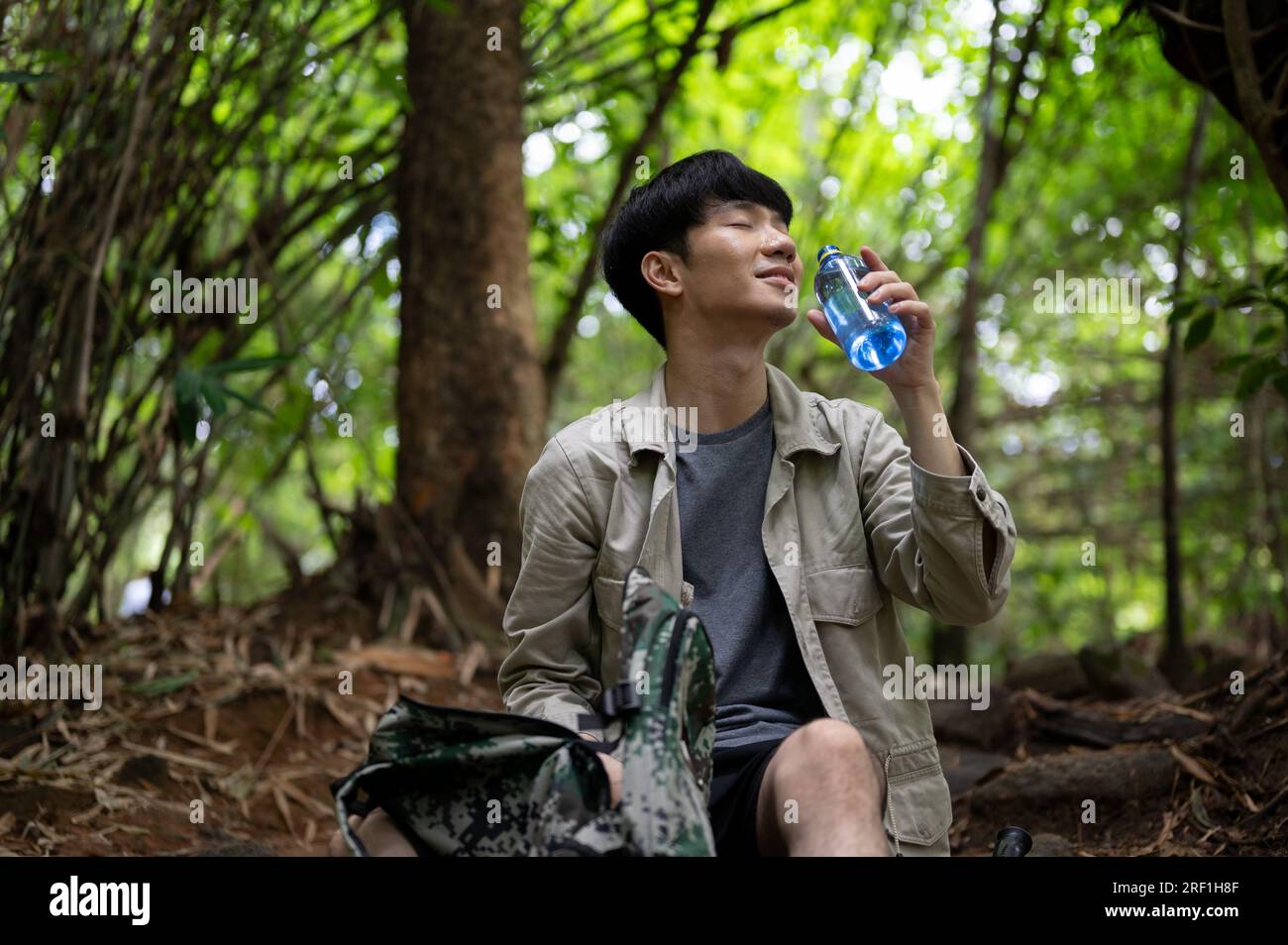 Ein entspannter und glücklicher männlicher asiatischer Wanderer oder Trekker sitzt, trinkt Wasser und macht eine kleine Pause während seiner Wanderung im grünen Wald. Stockfoto