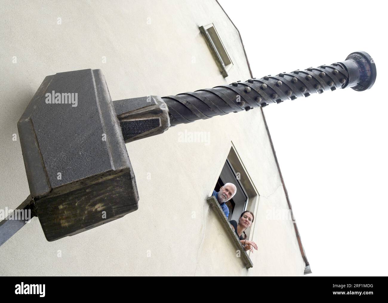 27. Juli 2023, Sachsen, Hohenprießnitz: Hinter dem Emblem ihrer Schmiede, einem 2,30 Meter langen Thors Hammer mit einem Gewicht von etwa 350 Kilogramm, der an der Wand des Hauses befestigt ist, schauen Schmied Marika Widdermann (r) und ihr Vater, Roger Widdermann, Maschinenbauingenieur und Schlosser von Ausbildung, aus einem Fenster. Das Vater-Tochter-Team mit seinem Schmied ist eines der wenigen Handwerksbetriebe, die dieses uralte Handwerk in Deutschland noch praktizieren. Neben individuellen Utensilien und Schmuck stellen sie auch geschmiedete Möbel für Kunden im in- und Ausland sowie Feuerschüsseln und Feuerbälle her Stockfoto