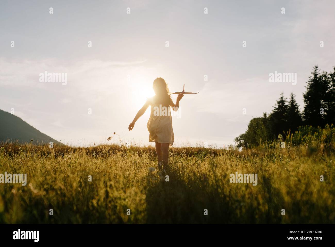 Silhouette eines verspielten Kindes, das mit dem Flugzeug im Hintergrund läuft, atemberaubender sommerlicher, warmer Sonnenuntergang und majestätische Berge. Das Konzept der Traumfreiheit. Kind Stockfoto