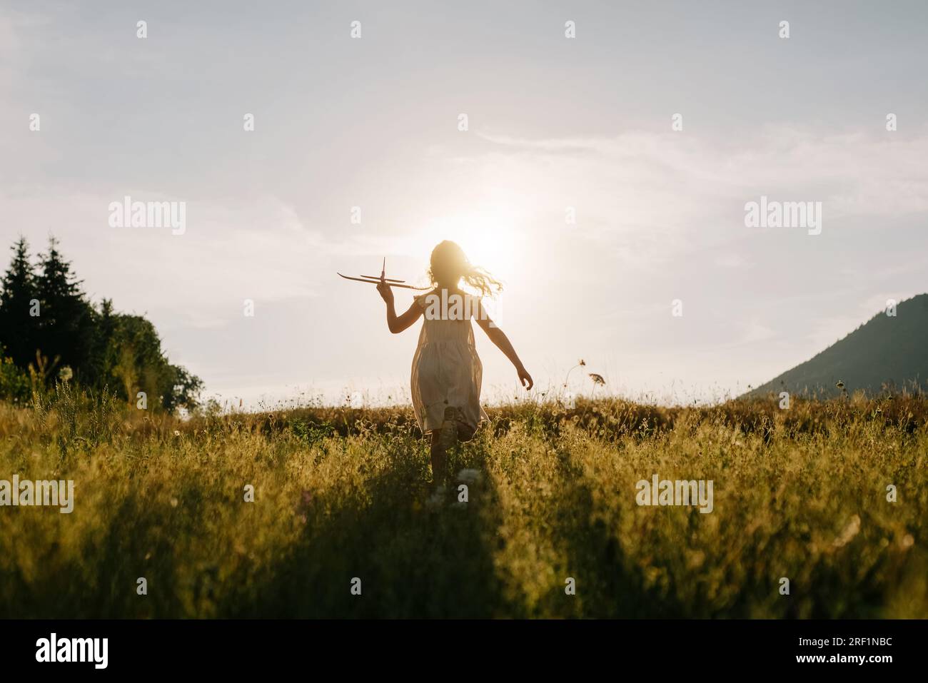 Silhouette eines jungen Mädchens, das mit dem Flugzeug auf majestätischen Bergen im Hintergrund bei warmem Sonnenuntergang im Sommer läuft. Das Konzept der Traumfreiheit. Spielerisches Kind rennt Stockfoto
