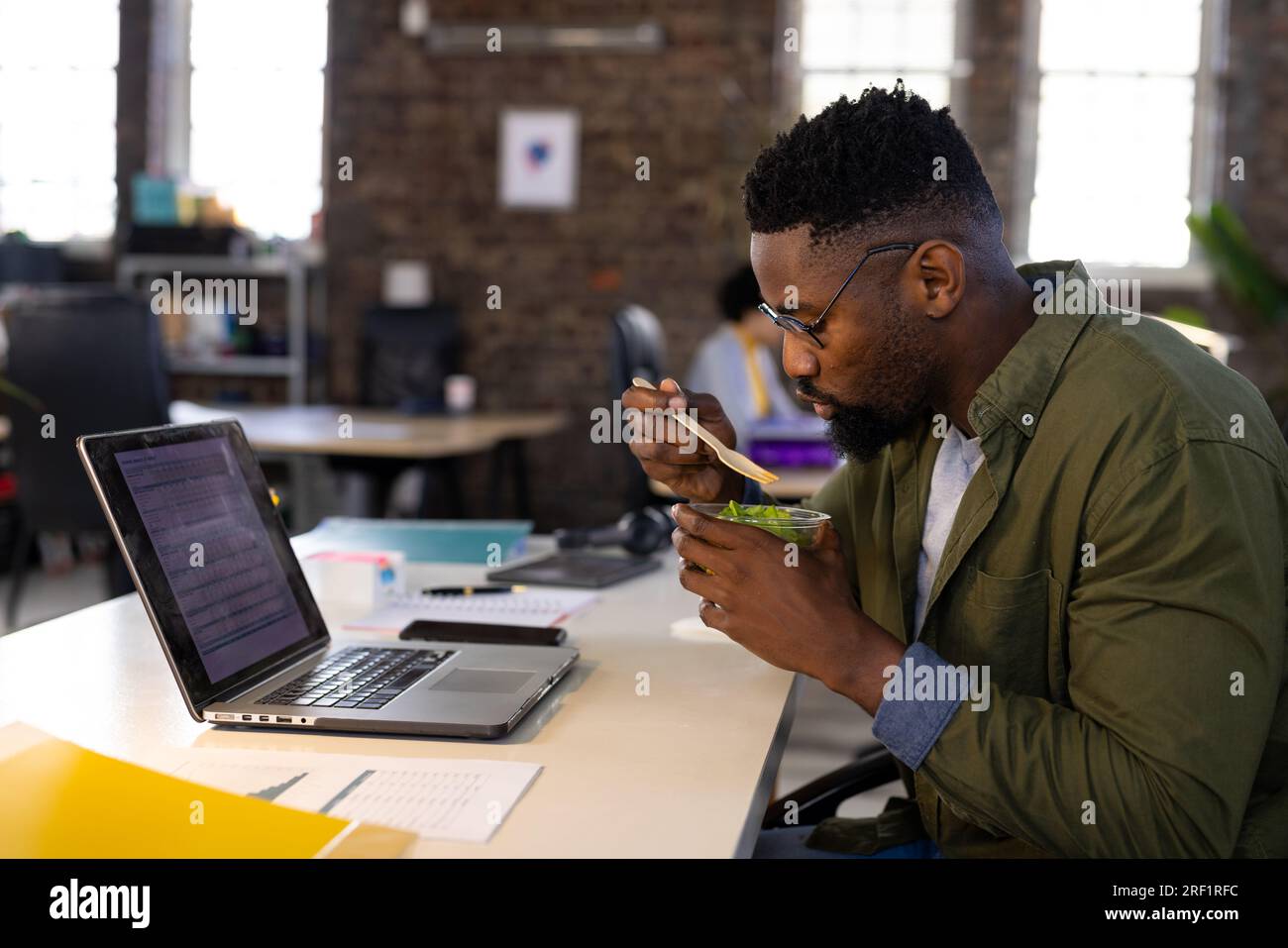 Ein afroamerikanischer Geschäftsmann, der ein Notebook benutzt und Salat zum Mitnehmen am Schreibtisch im Büro isst Stockfoto