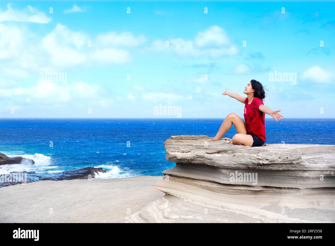 Junge Frau, die auf einer großen Felsenklippe am hawaiianischen Ozean sitzt, Arme hoch und die Brise genießt Stockfoto