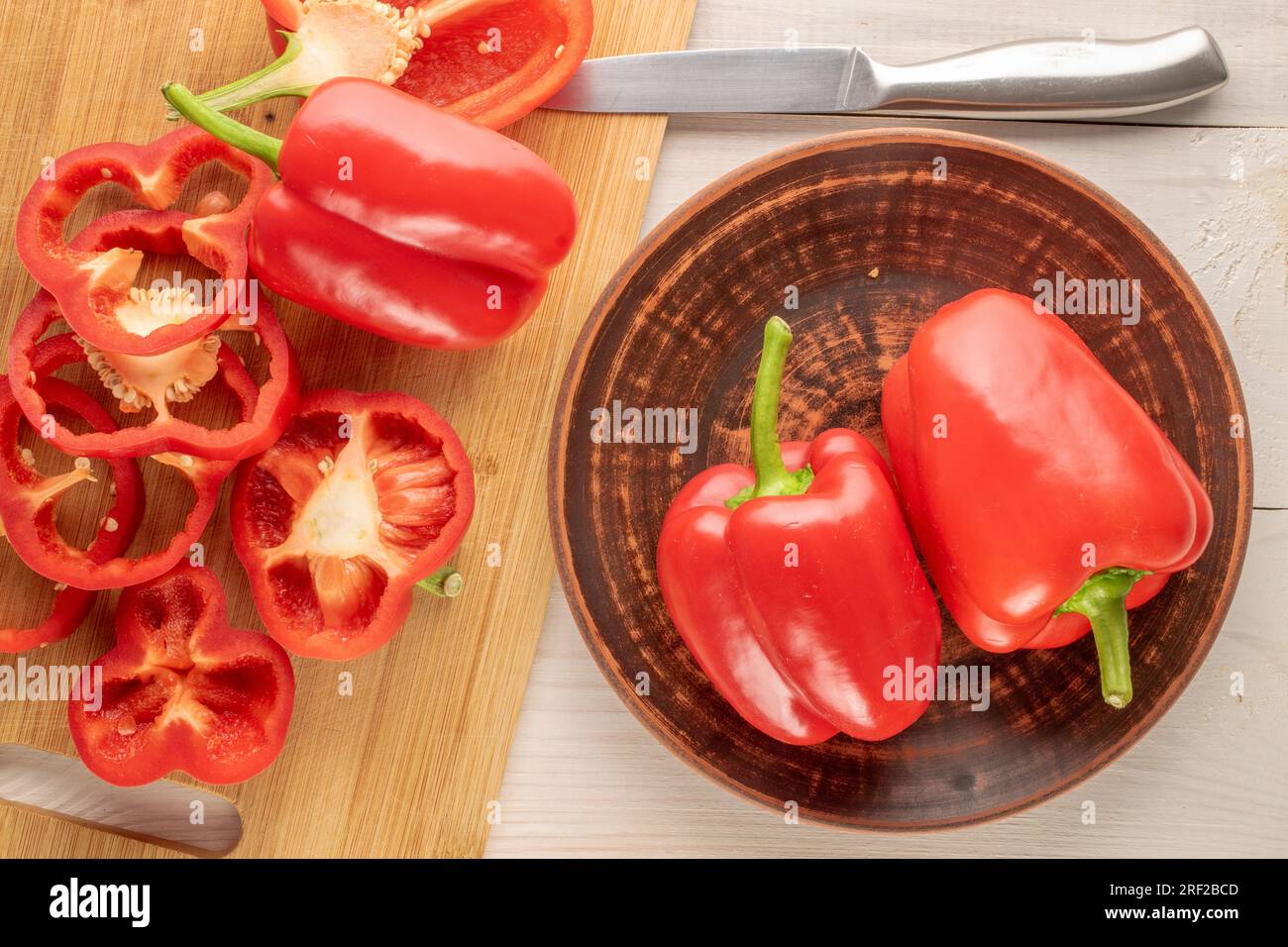 Mehrere rote Paprika im Ganzen und in Scheiben geschnitten mit Tonplatte und Messer auf Holztisch, Makro, Draufsicht. Stockfoto