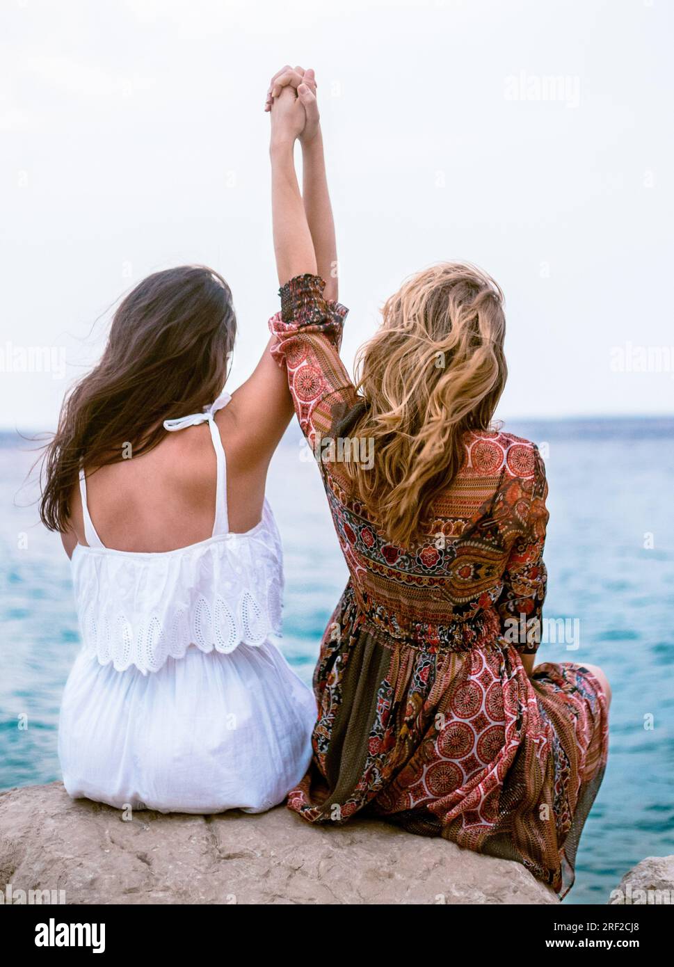 Rückansicht von zwei Frauen, die auf einem Felsen über dem Meer stehen und Hand halten Stockfoto