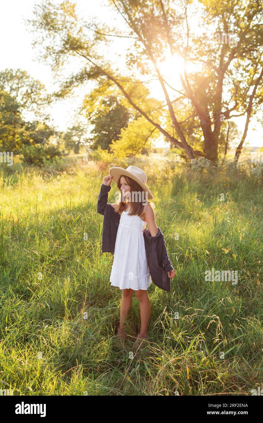 Ein kleines Mädchen mit Hut, das im hohen Gras steht und im Sommer den Sonnenuntergang erstrahlt Stockfoto