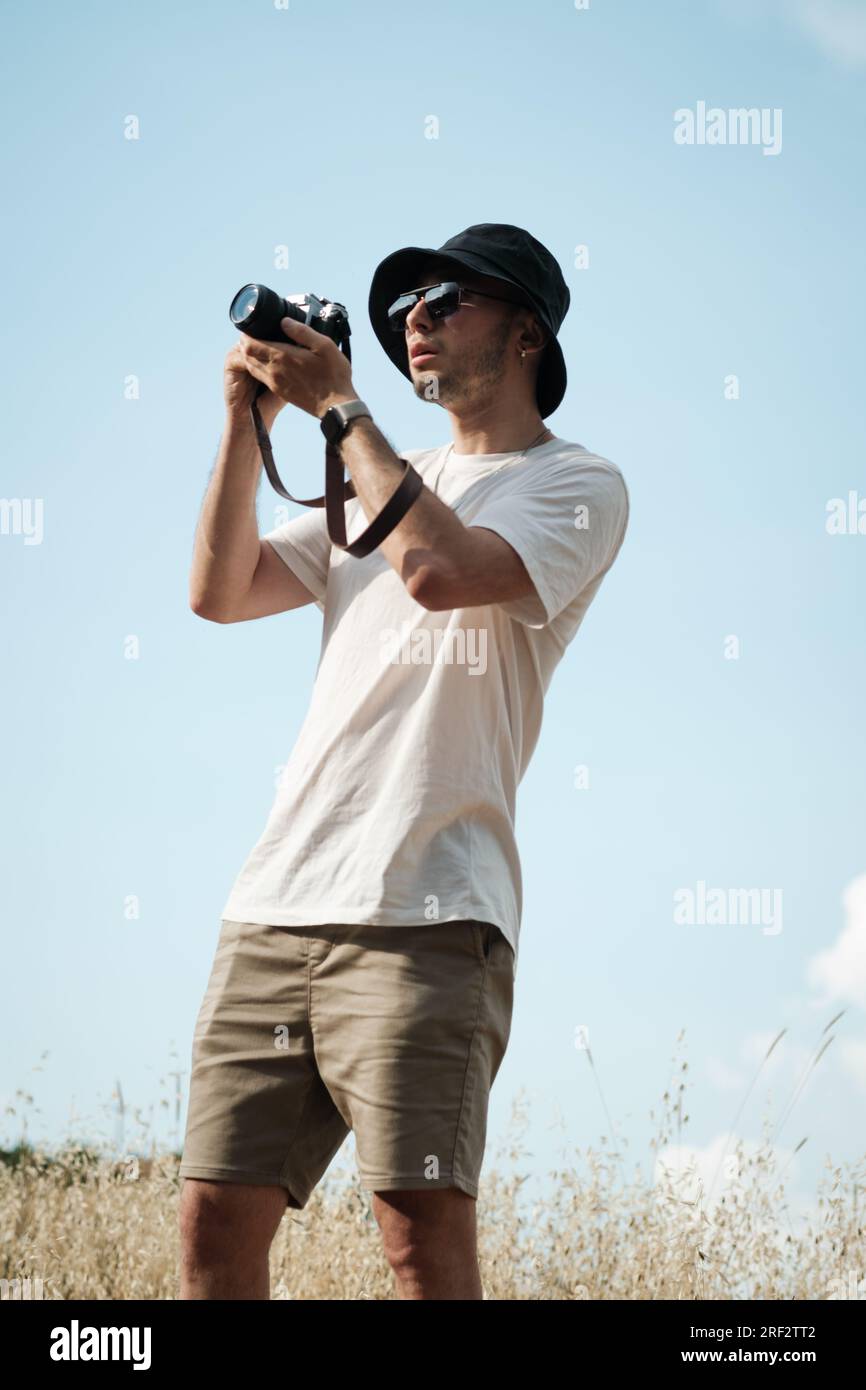 Ein Porträt eines Mannes in seinen Zwanzigern, lässig gekleidet in einem weißen T-Shirt, beigen Shorts, Sonnenbrille und einer schwarzen Seemannsmütze, mit einem klassischen Kameraschlub Stockfoto