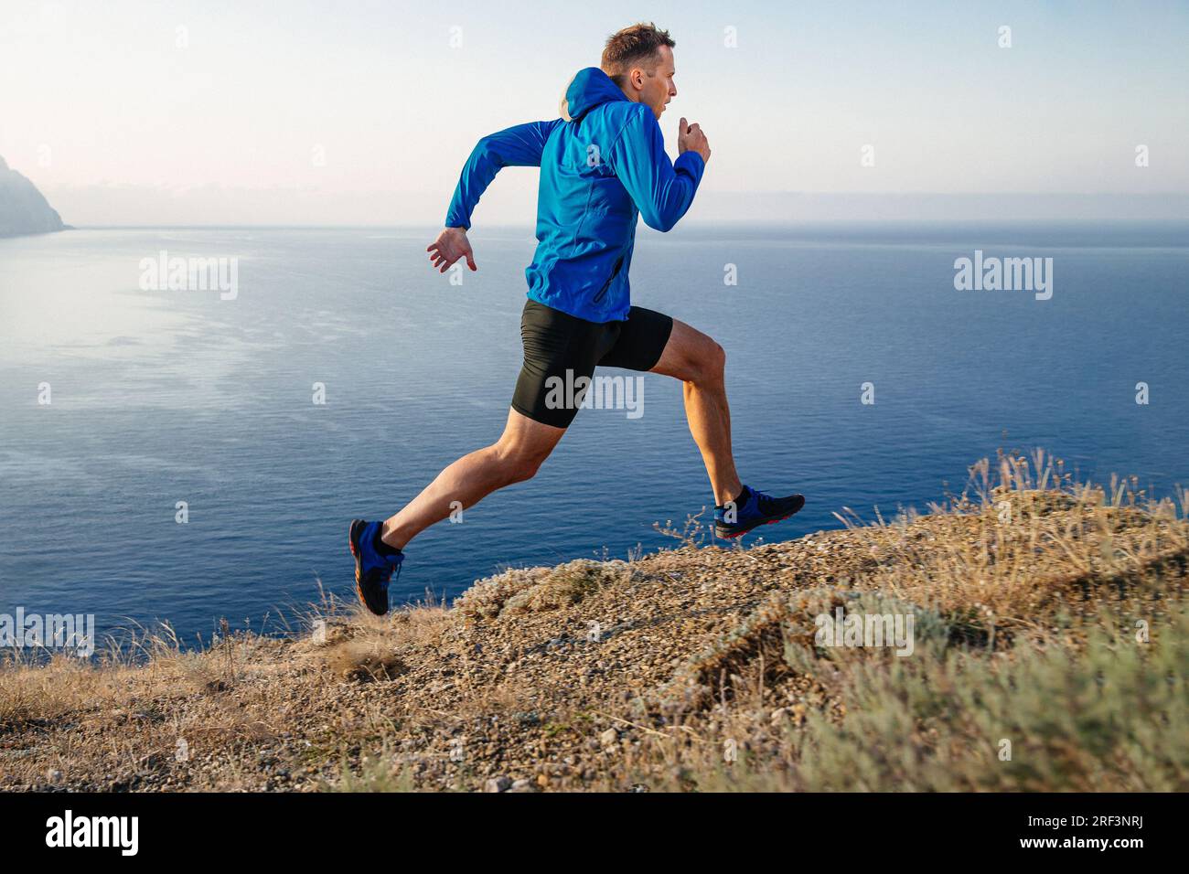 Männlicher Läufer läuft bergauf in blauer Jacke und schwarzen Strumpfhosen, Hintergrund von Himmel und Meer Stockfoto