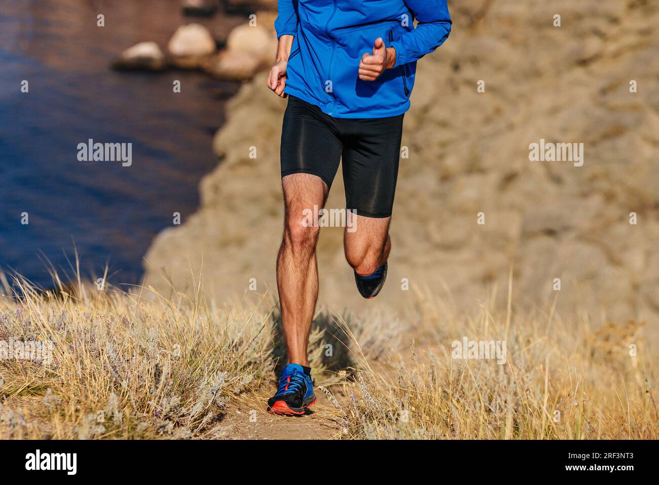 Läufer mit Vorderansicht läuft bergauf in blauer Jacke und schwarzen Strumpfhosen, auf trockenem Gras Stockfoto