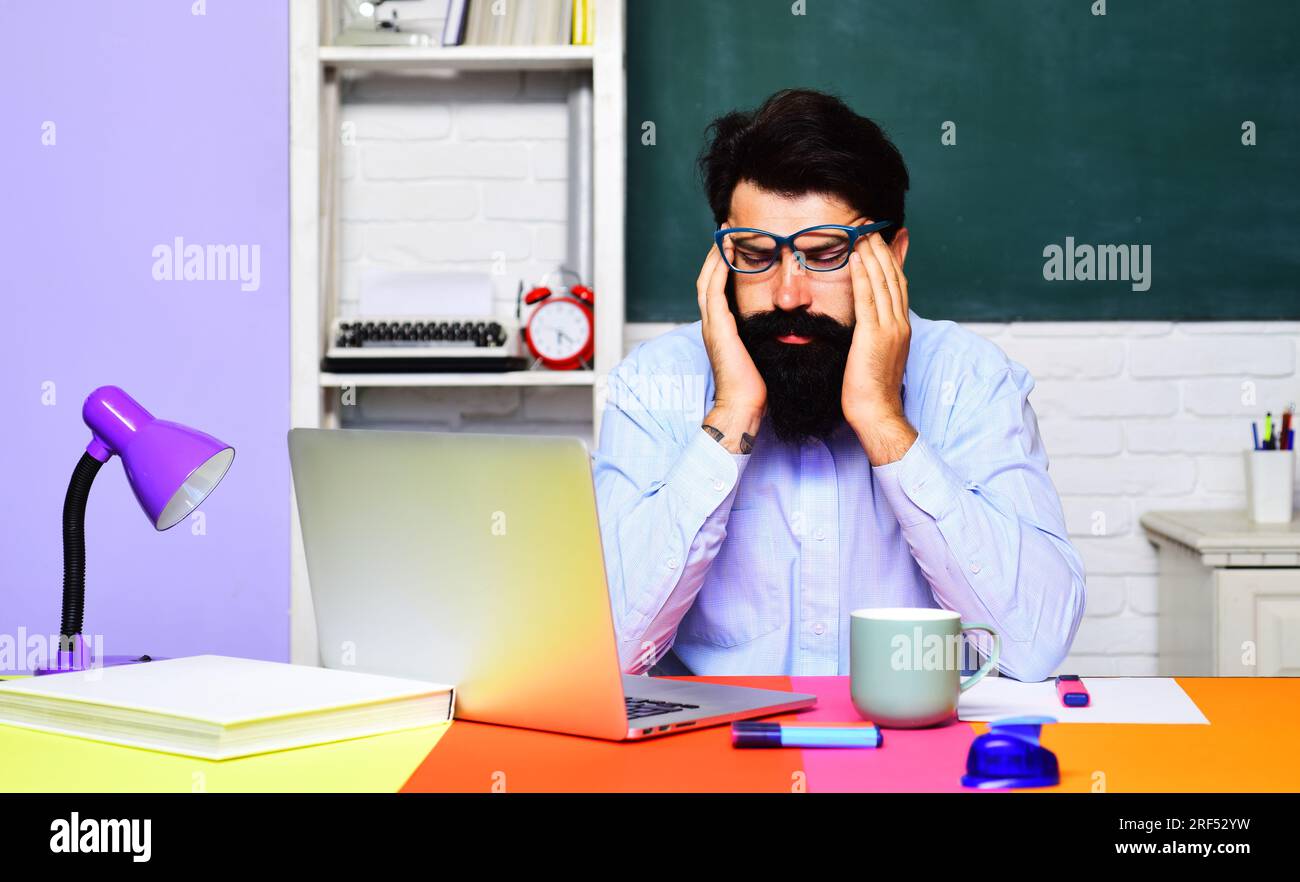 Erschöpfter gestresster Lehrer mit Kopfschmerzen am Schreibtisch im Klassenzimmer. Müde männliche Schülerin, die sich nach einer riesigen Menge Computer Migräne fühlt Stockfoto