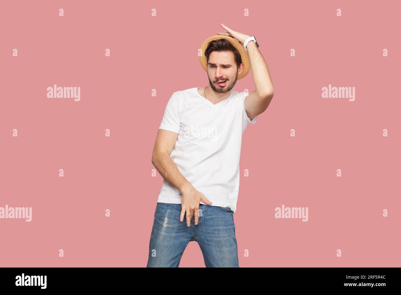 Porträt eines aufgeregten, verspielten, gutaussehenden Mannes mit weißem T-Shirt und Hut, der lustige Posen hat und Zeit auf einer Party verbringt. Studio-Aufnahme im Innenbereich isoliert auf pinkfarbenem Hintergrund. Stockfoto