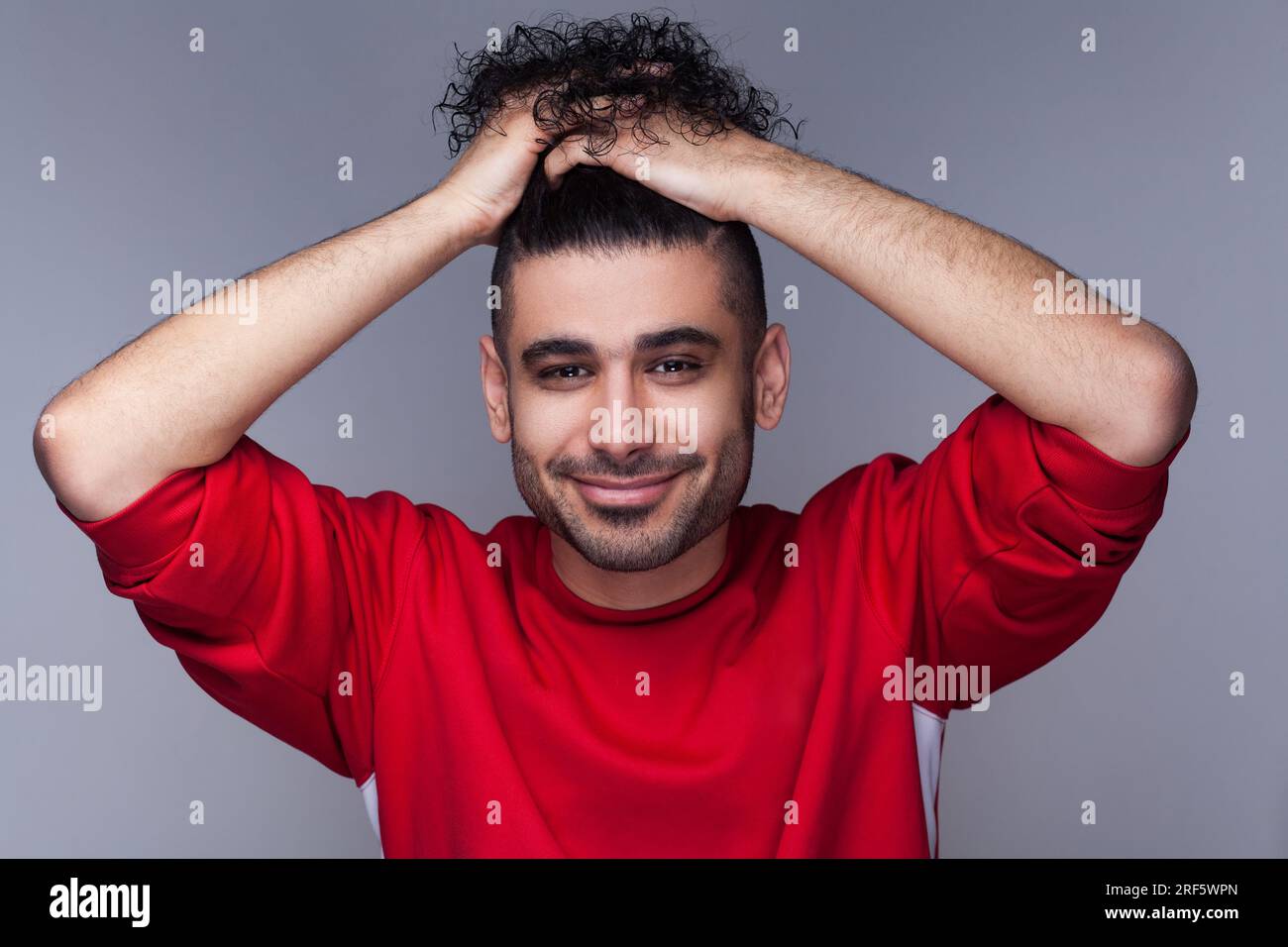 Porträt eines attraktiven, lächelnden, bärtigen Mannes mit lockigem Haar, der roten Pullover trägt, in die Kamera schaut, sein Haar sammelte. Studioaufnahmen im Innenbereich isoliert auf grauem Hintergrund. Stockfoto