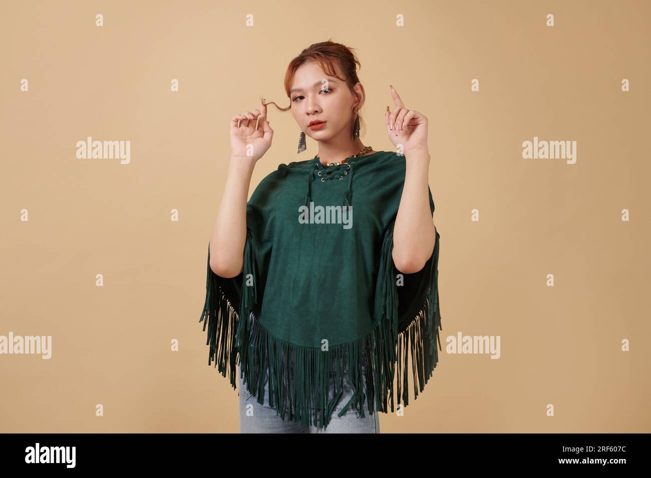 Porträt einer jungen Transgender-Frau im grünen Poncho, die ihr Haar berührt und in die Kamera schaut Stockfoto