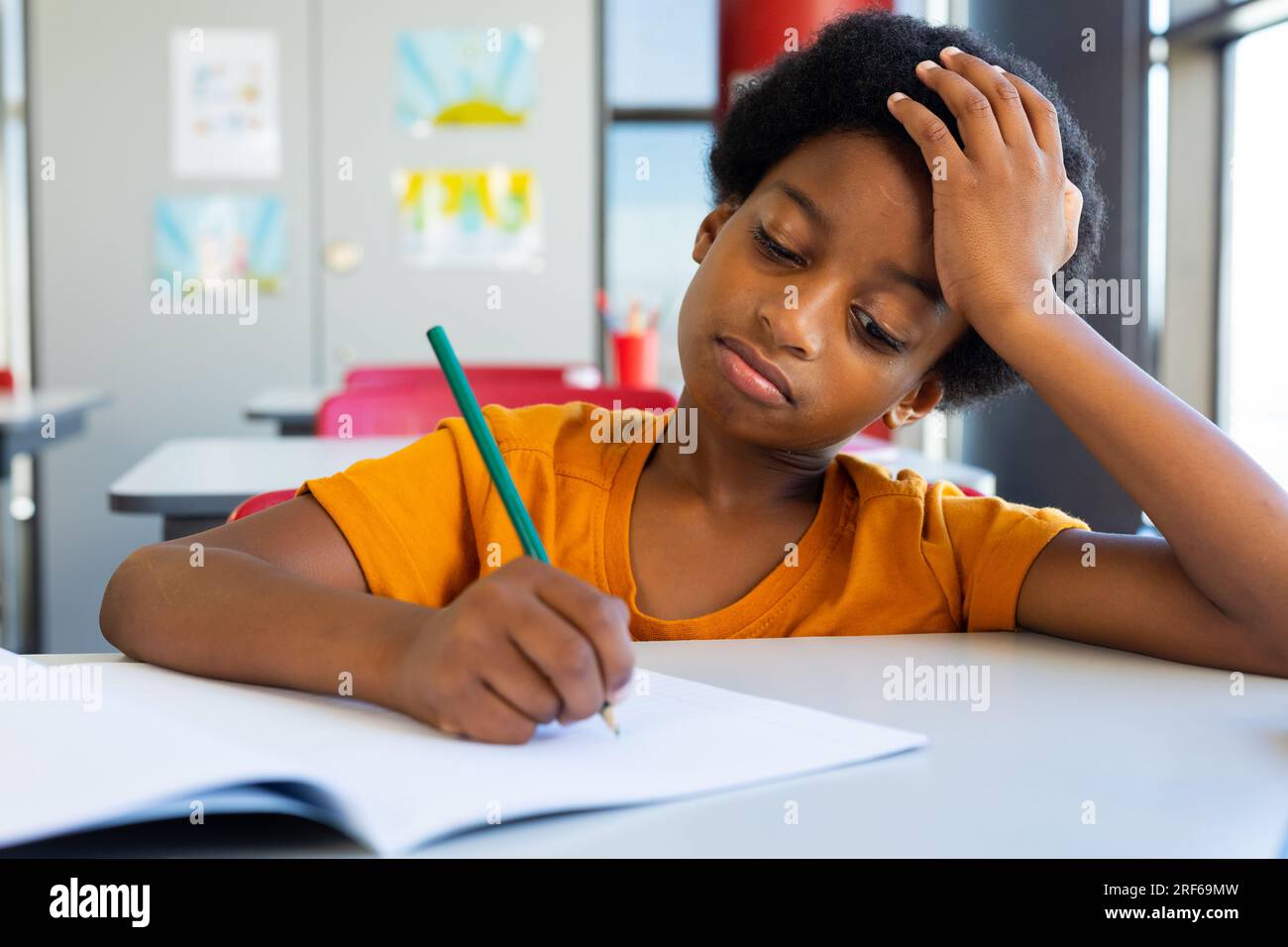 Erschöpfter, birassistischer Schuljunge, der am Schreibtisch sitzt und im Klassenzimmer schreibt Stockfoto