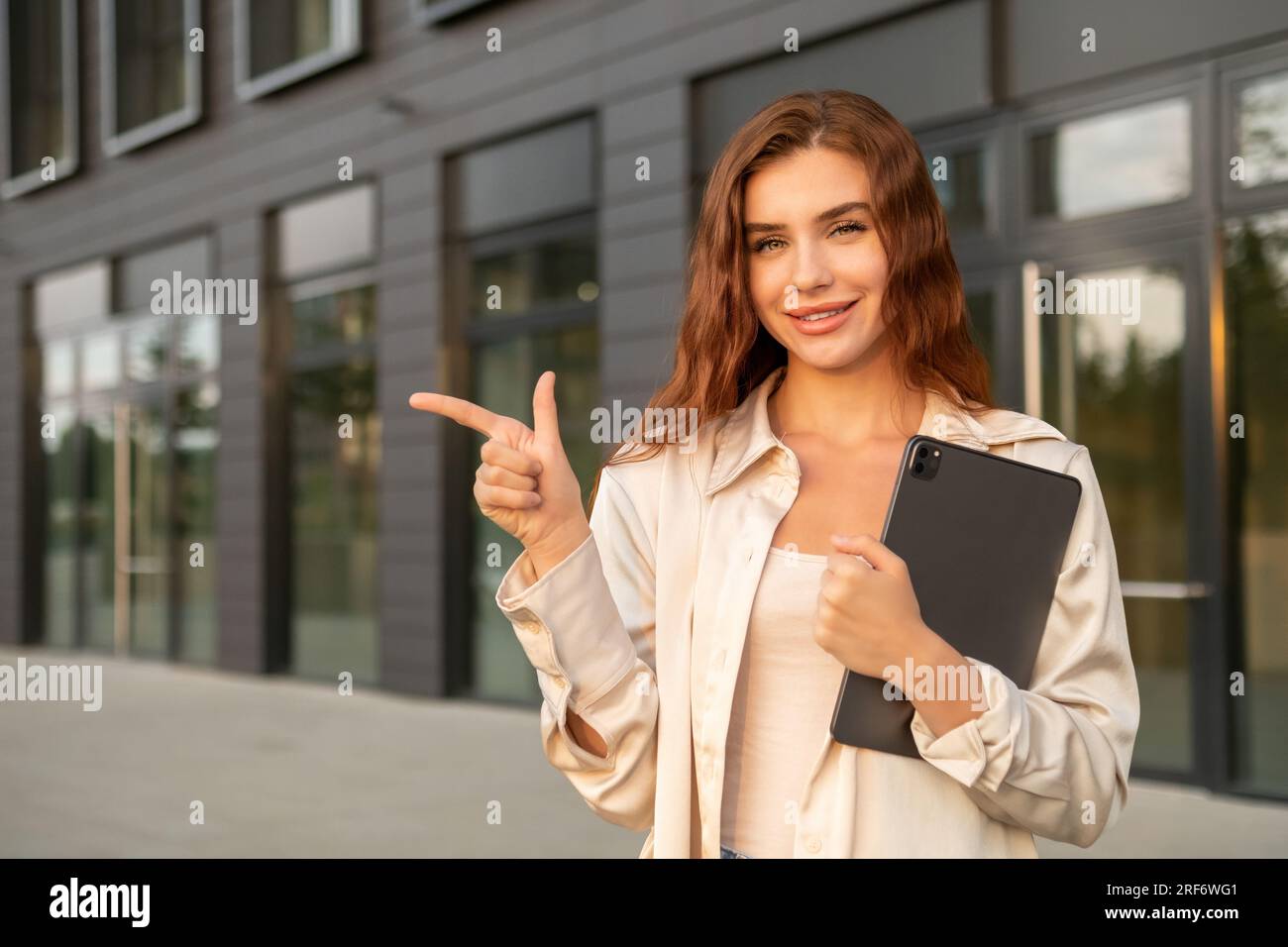 Lächelnde, rothaarige Geschäftsfrau, die ein Tablet hielt und nach links zeigte, in der Nähe des Business Centers. Stockfoto