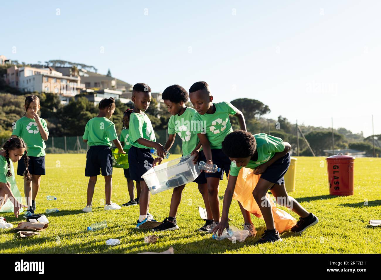 Glückliche, vielfältige Kinder, die Plastikabfälle auf dem sonnigen Sportplatz der Grundschule sammeln Stockfoto