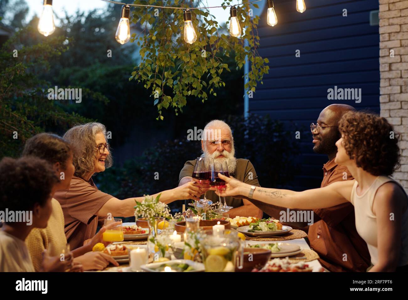 Seitenansicht einer großen interkulturellen sechsköpfigen Familie mit Gläsern Rotwein auf dem Tisch, serviert mit hausgemachten Speisen während des Abendessens im Freien Stockfoto