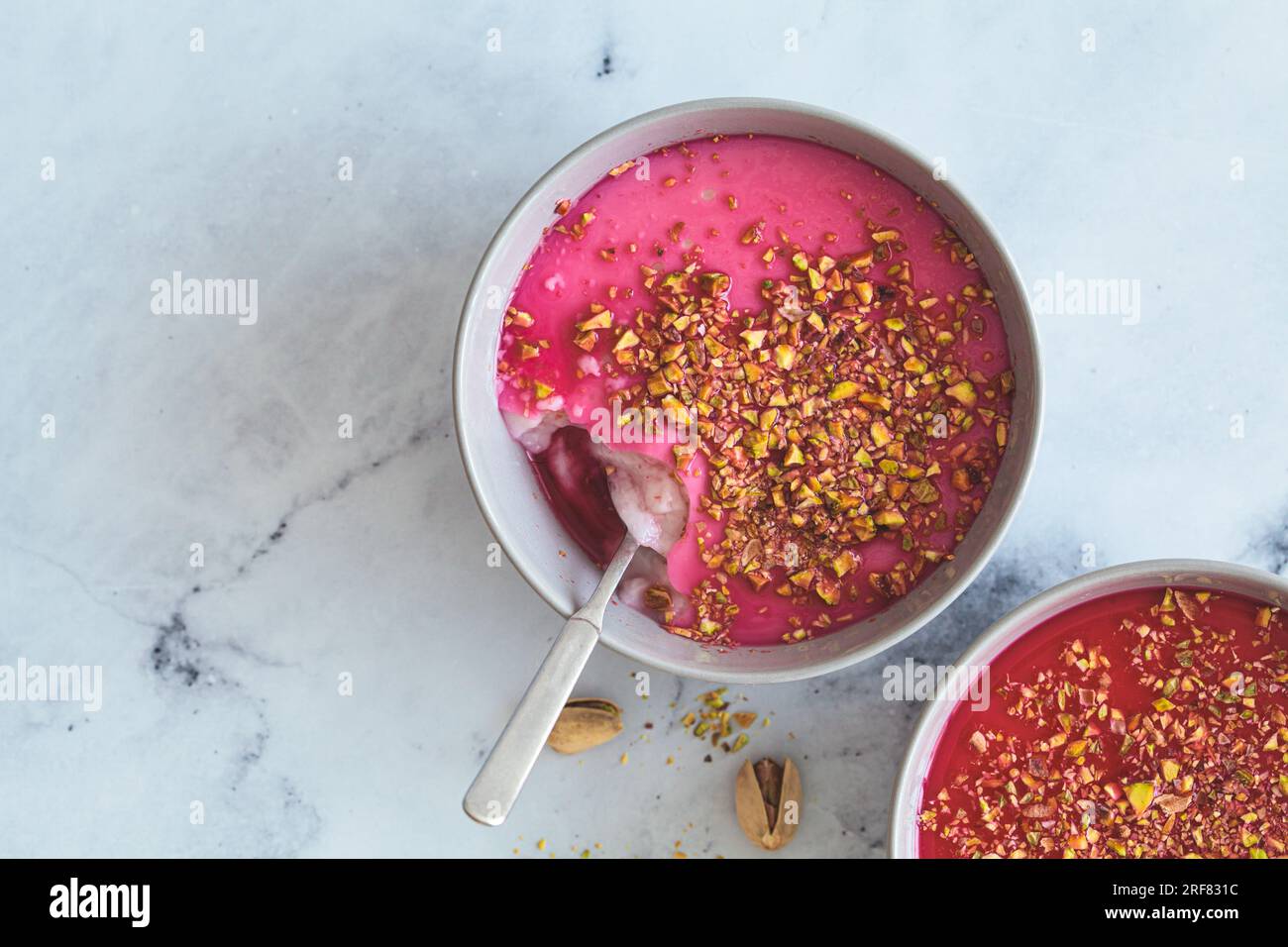 Malabi traditionelles arabisches Dessert. Milchpudding mit rosafarbenem Sirup und Pistazien in grauer Schüssel, Draufsicht auf weißem Marmorhintergrund. Stockfoto