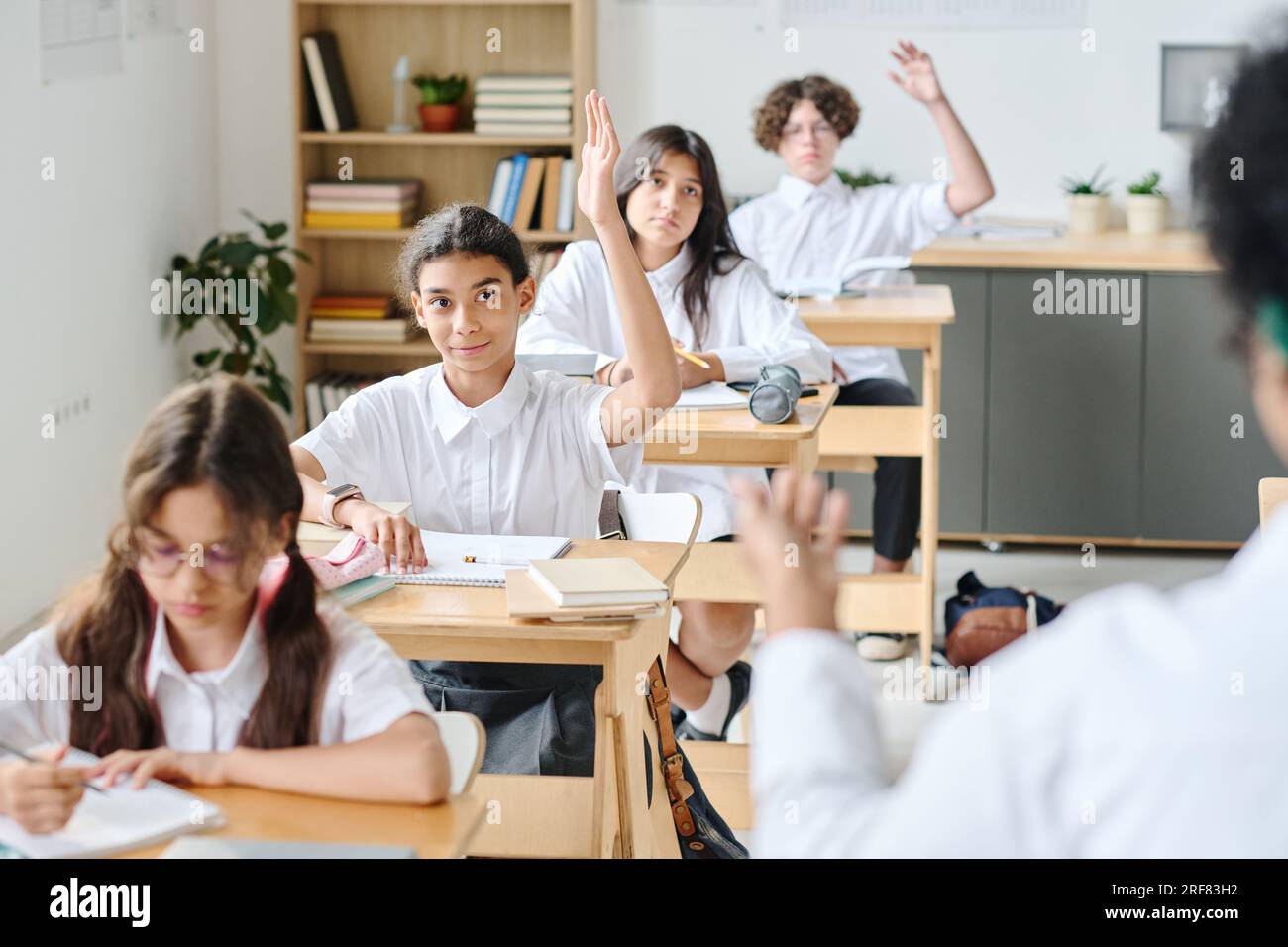 Schulkinder sitzen an Schreibtischen und heben die Hand, um die Frage des Lehrers zu beantworten Stockfoto