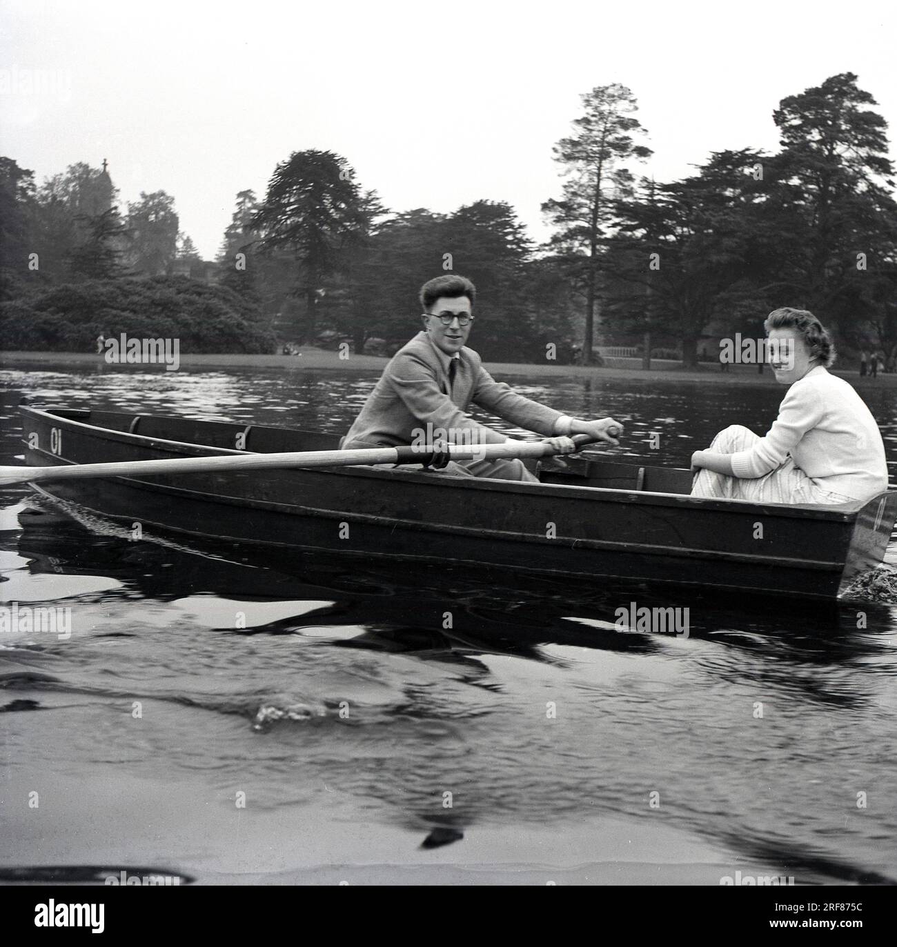 1950er, historisch, draußen an einem See, ein junges Paar, das zusammen in einem Ruderboot sitzt, der Mann, der einen Anzug und eine Krawatte trägt, mit den Rudern in der Hand, England, Großbritannien. Stockfoto
