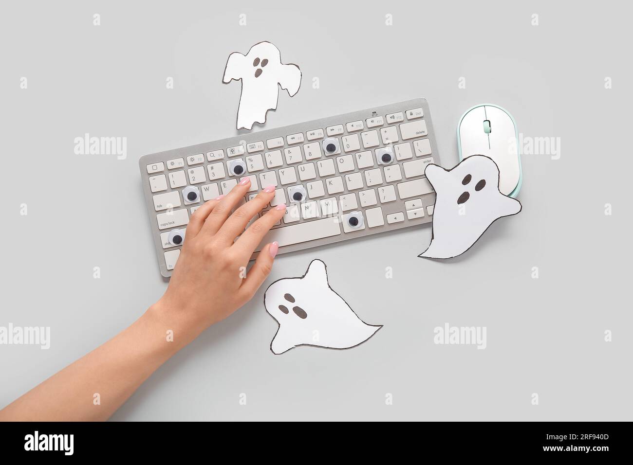 Weibliche Hand mit moderner Computertastatur, Maus und Papiergeistern für Halloween auf grauem Hintergrund Stockfoto