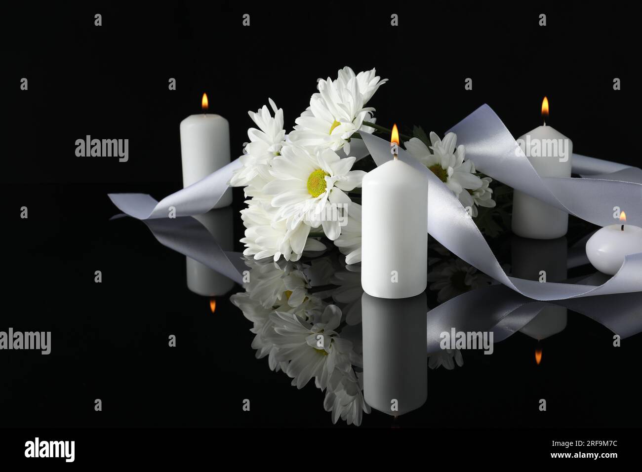 Weiße Chrysanthemen und brennende Kerzen auf schwarzer Spiegeloberfläche in Dunkelheit, Platz für Text. Begräbnissymbole Stockfoto