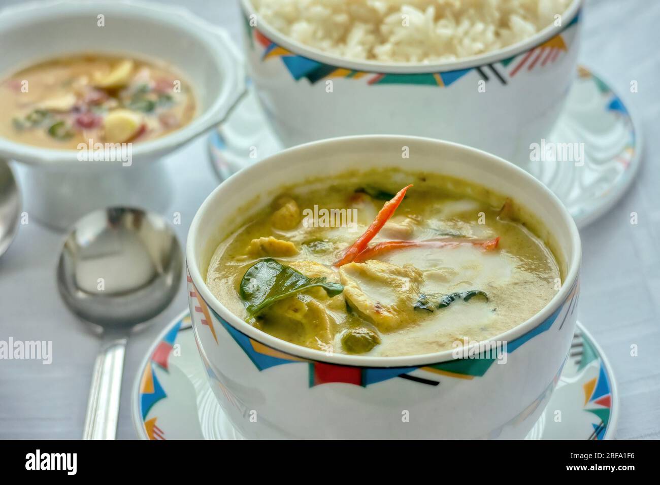 Eine Mahlzeit mit grünem thailändischem Curry-Huhn, serviert mit Reis in gemusterten Schüsseln im westlichen Stil auf einer weißen Tischdecke. Geringe Schärfentiefe. Stockfoto