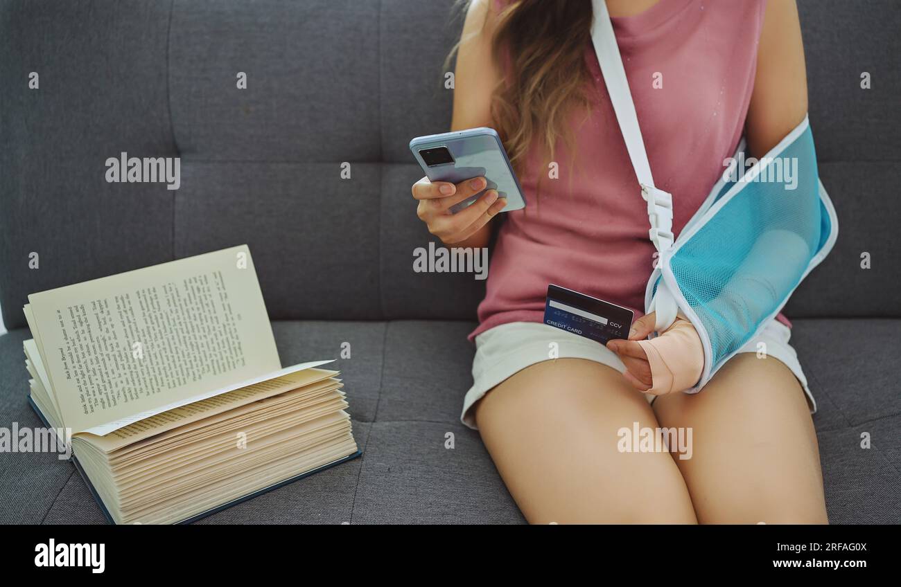 Junge asiatische Frau, die aufgrund von Wunde Arme eine weiche Schiene trägt, mit Telefon und Kreditkarte auf dem Sofa zu Hause. Stockfoto