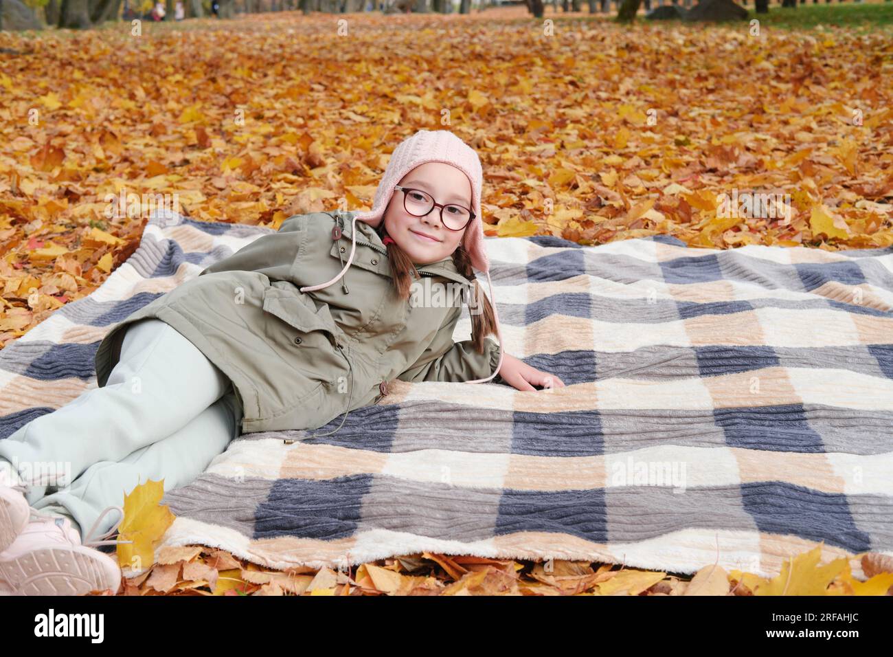 Mädchen in Herbstjacke, rosa Hut und Brille liegen auf einer Decke in einem Herbstpark. Das Mädchen lächelt und schaut in die Kamera. Horizontales Foto Stockfoto