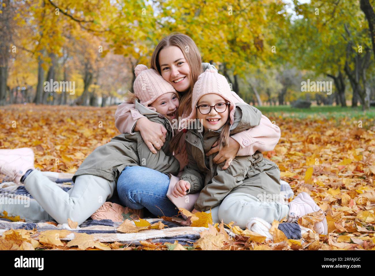 Eine glückliche Mutter mit ihren Zwillingstochter verbringt ihre Zeit damit, auf einer Decke in einem Herbstpark zu sitzen. Mutter umarmte ihre Töchter, lächelte und schaute auf die Ca Stockfoto